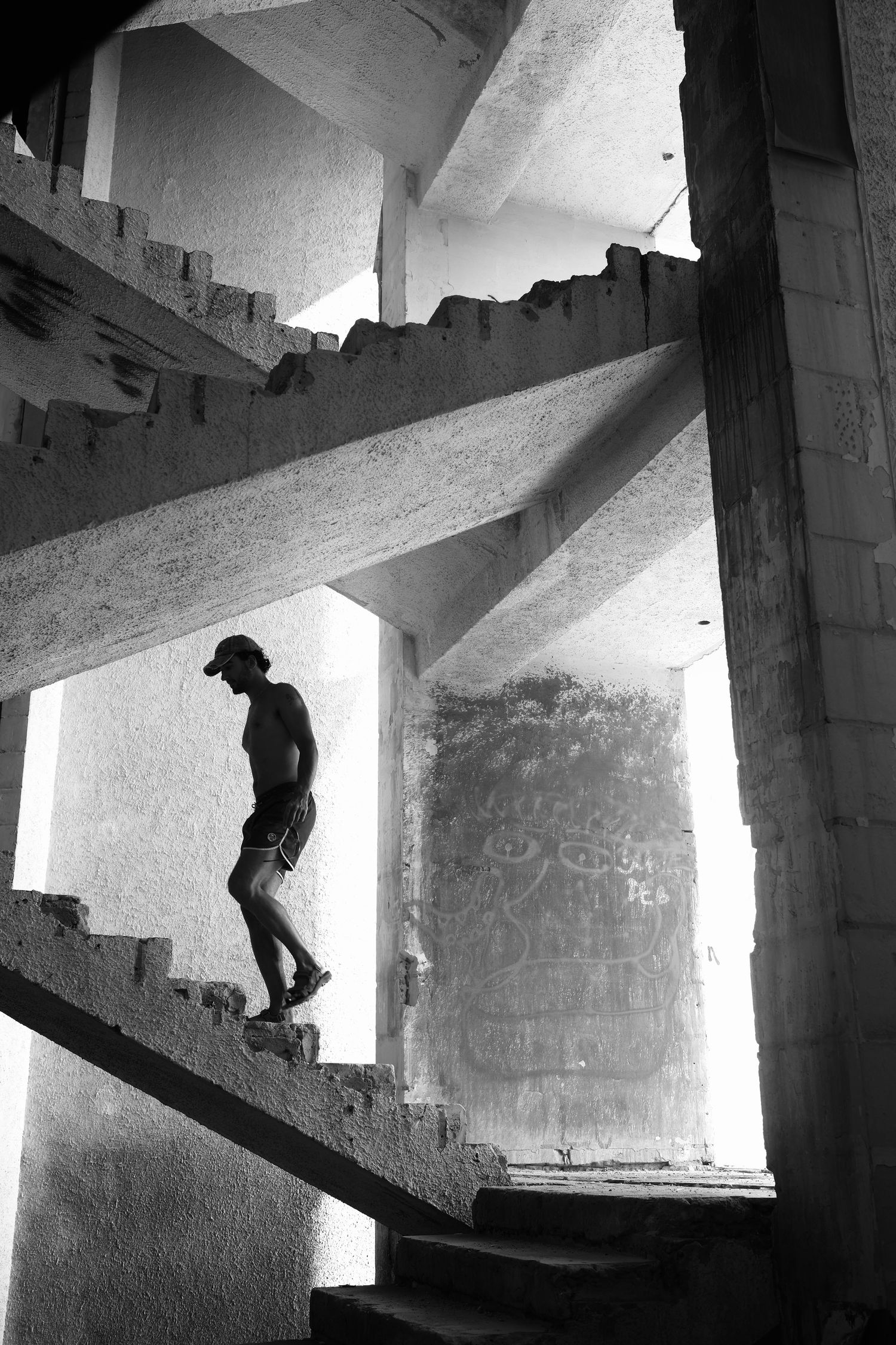 Black and white photo of shirtless man climbing concrete stairs in an abandoned building with graffiti walls.