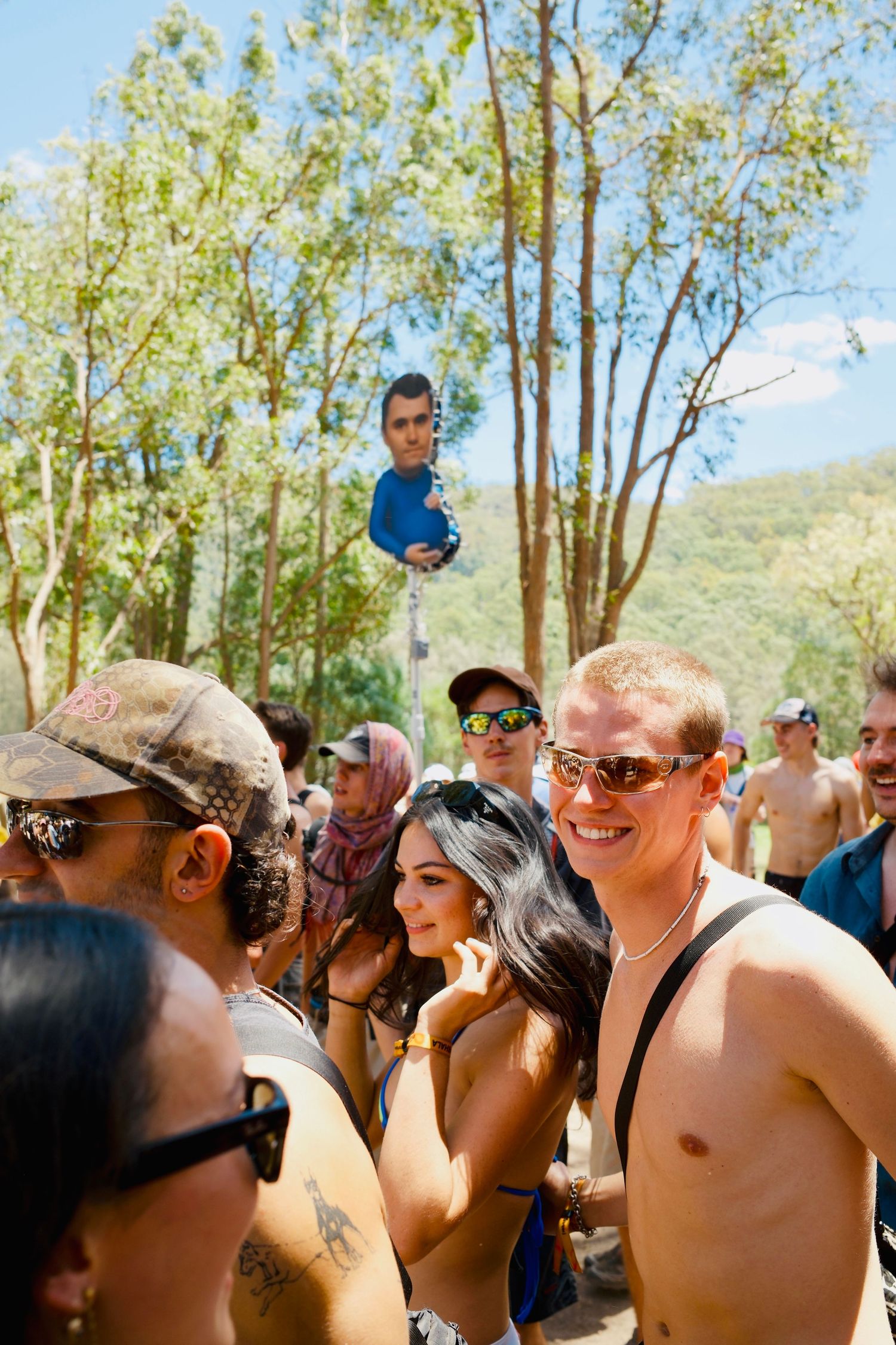 Smiling shirtless man and friends enjoying a sunny outdoor music festival crowd.