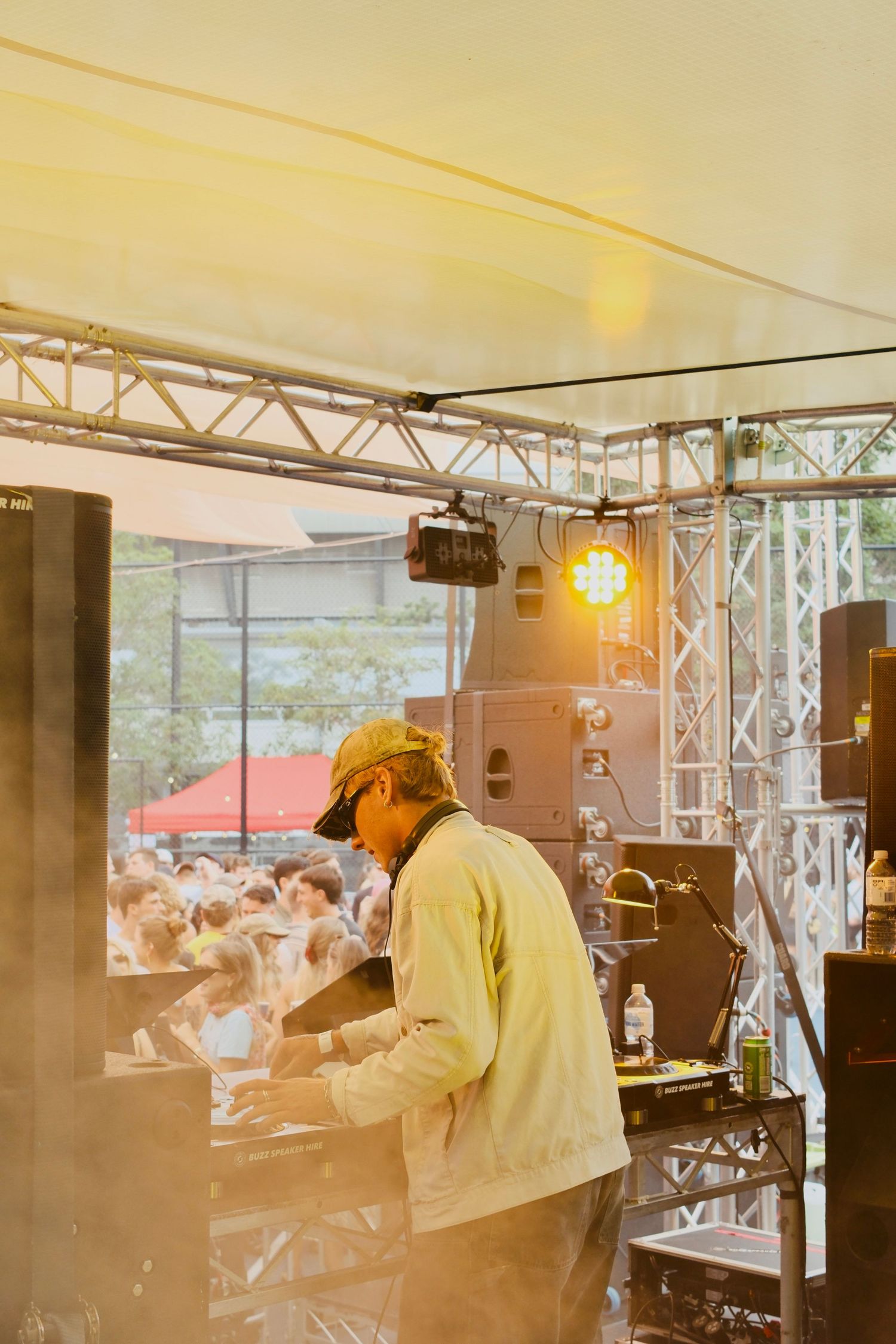 DJ performing on stage under bright lights at an outdoor music festival with a large crowd in the background.