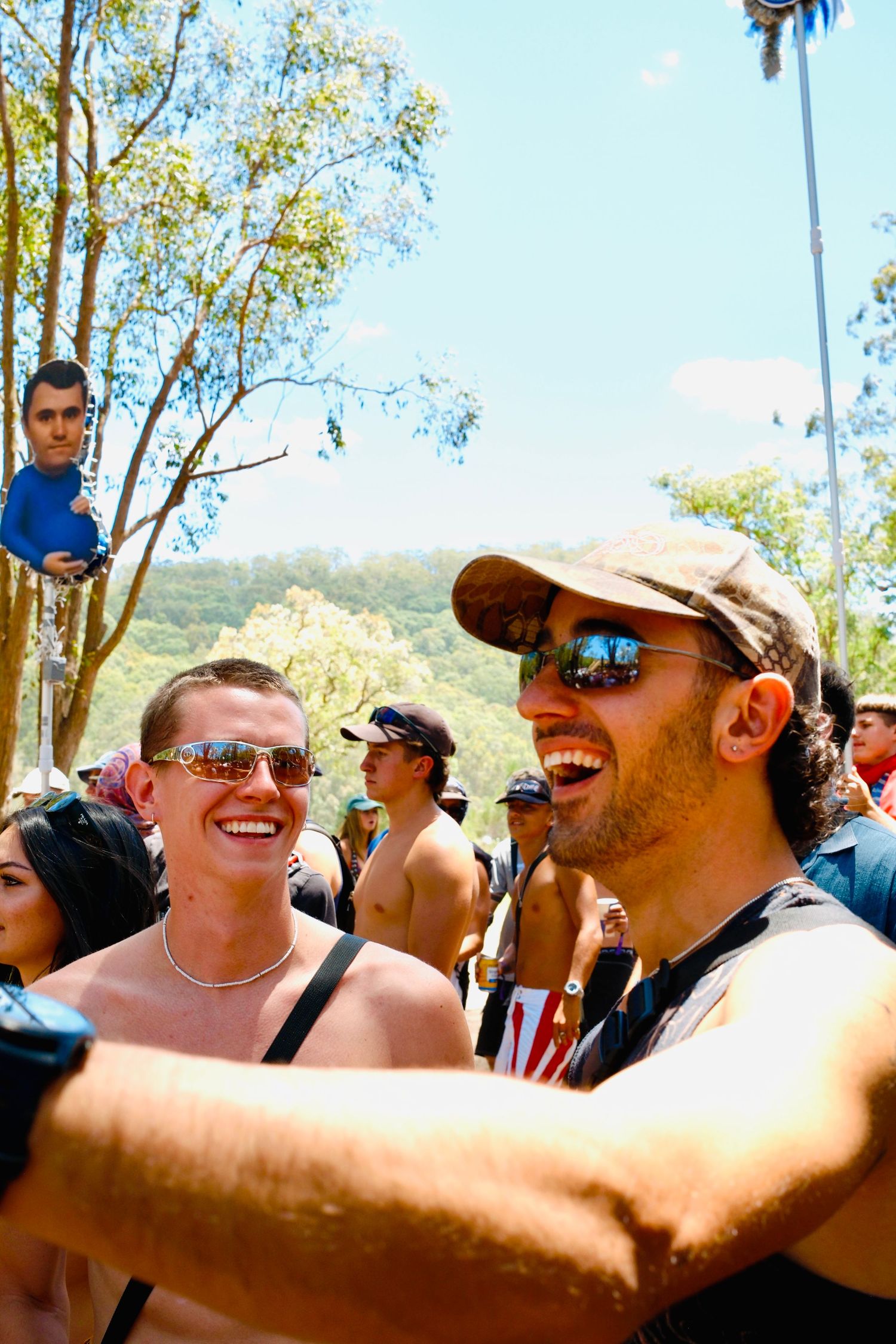 Two laughing men wearing sunglasses enjoying an outdoor music festival on a sunny day.