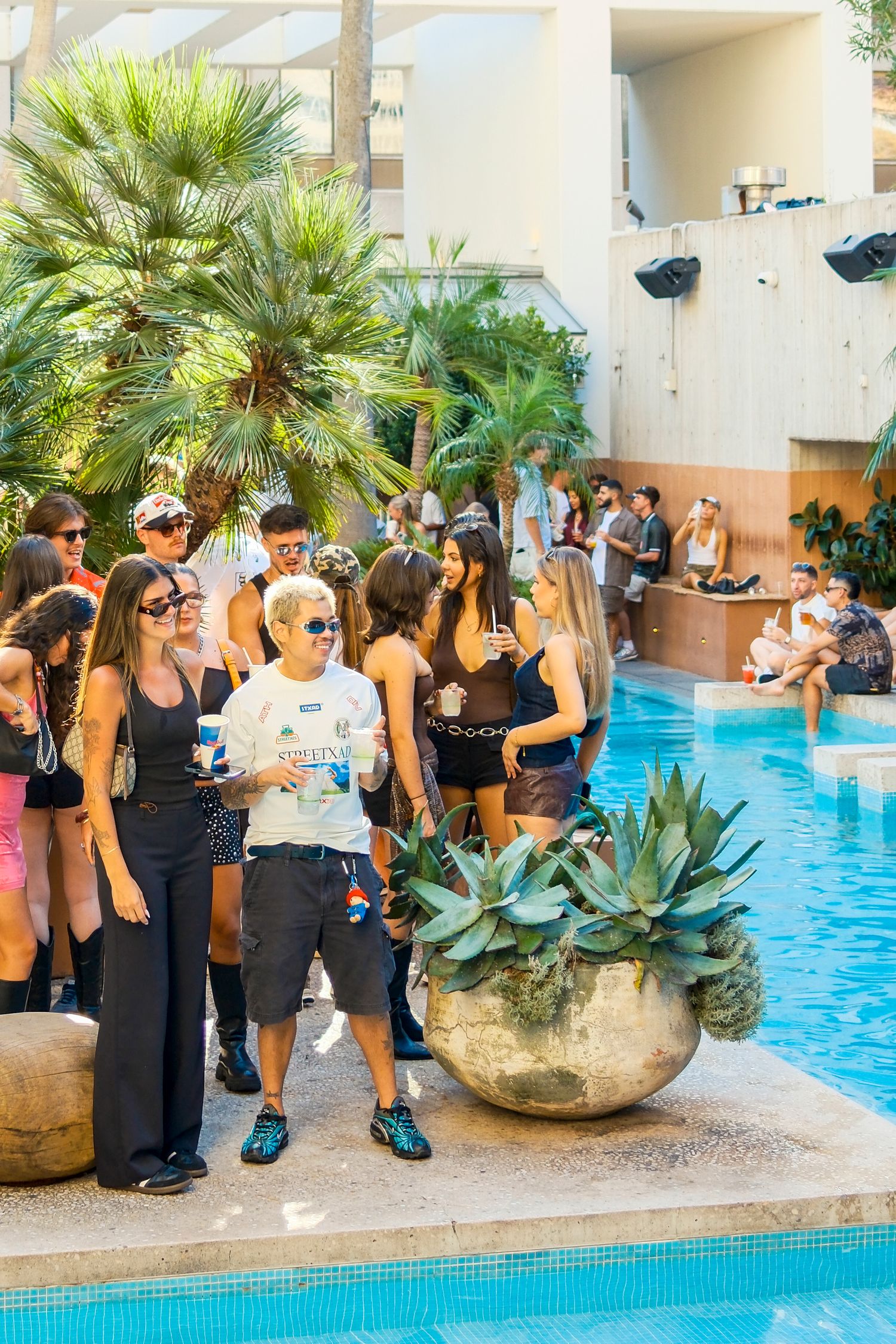 Guests mingle poolside at a luxury resort party, surrounded by palm trees, blue water, and tropical plants.