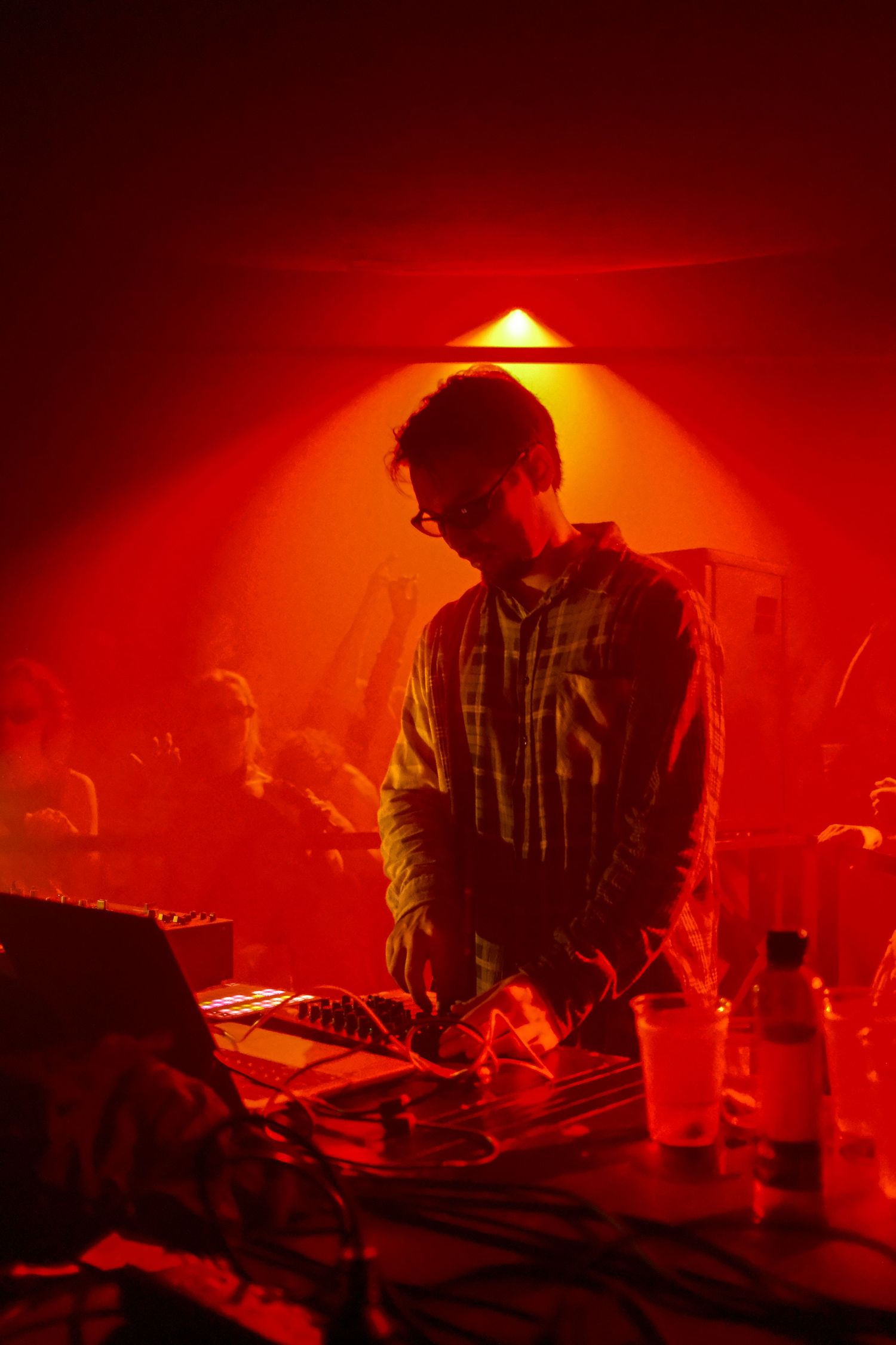 DJ performing under red atmospheric lighting at a live music event with crowd in background.