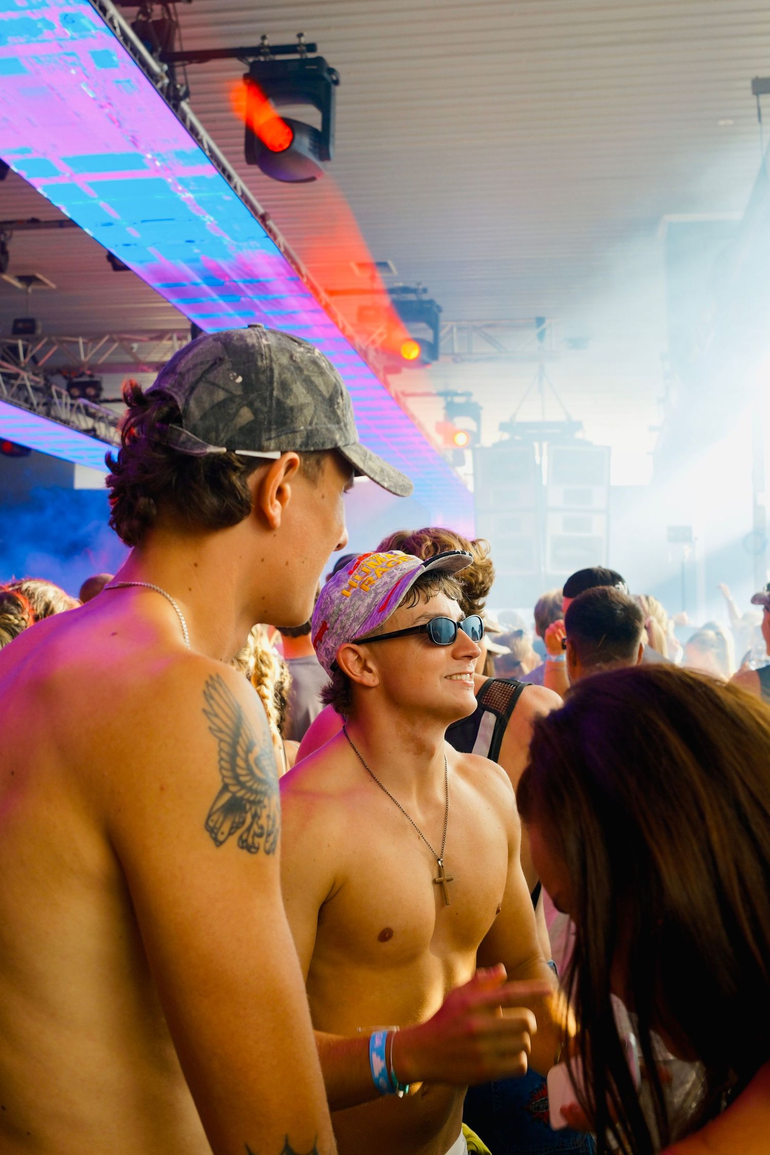 Shirtless tattooed men with caps dancing under colorful neon lights at an indoor music festival rave.
