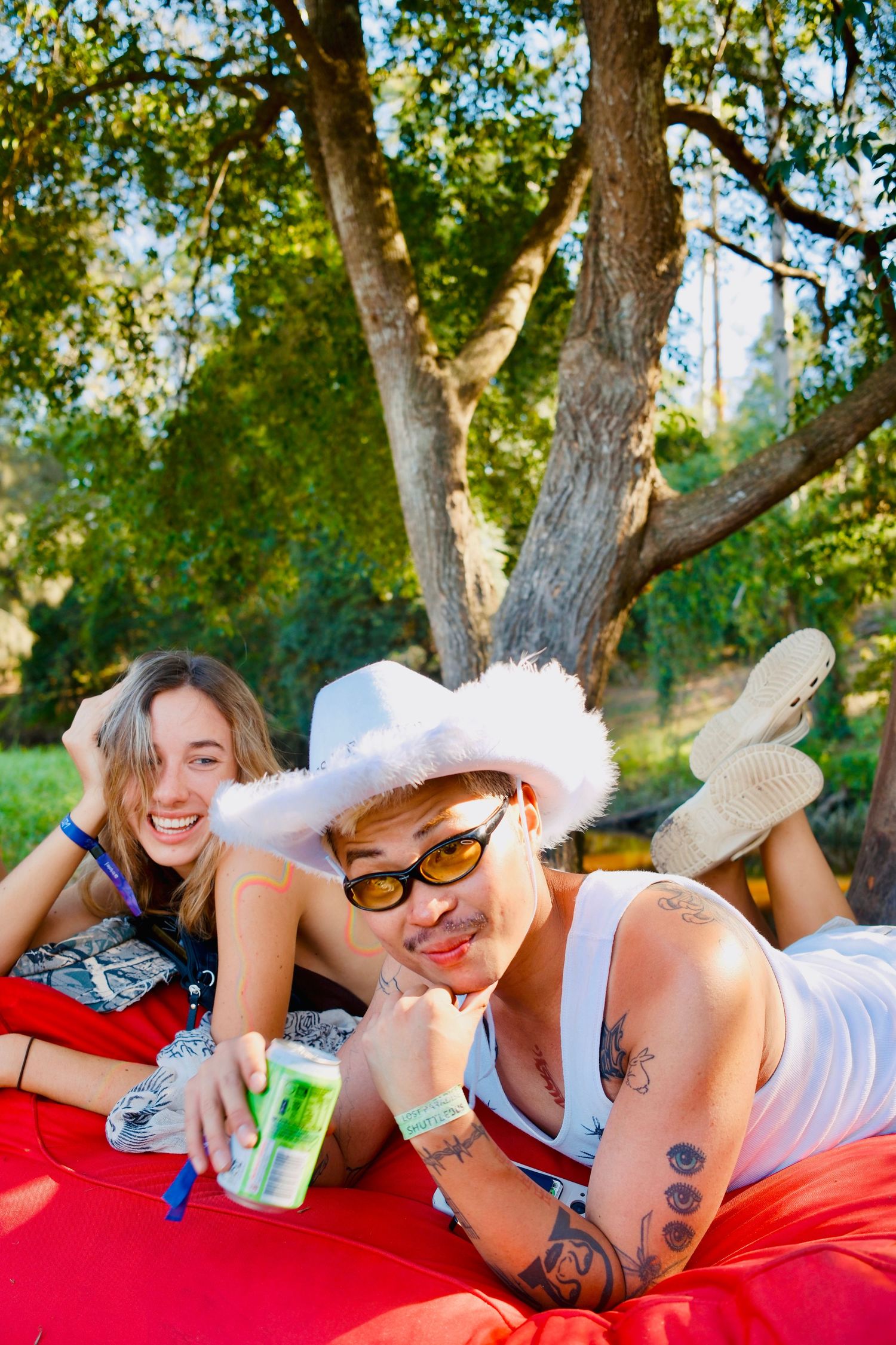 Two friends relaxing on red bean bags under a large tree at an outdoor festival.