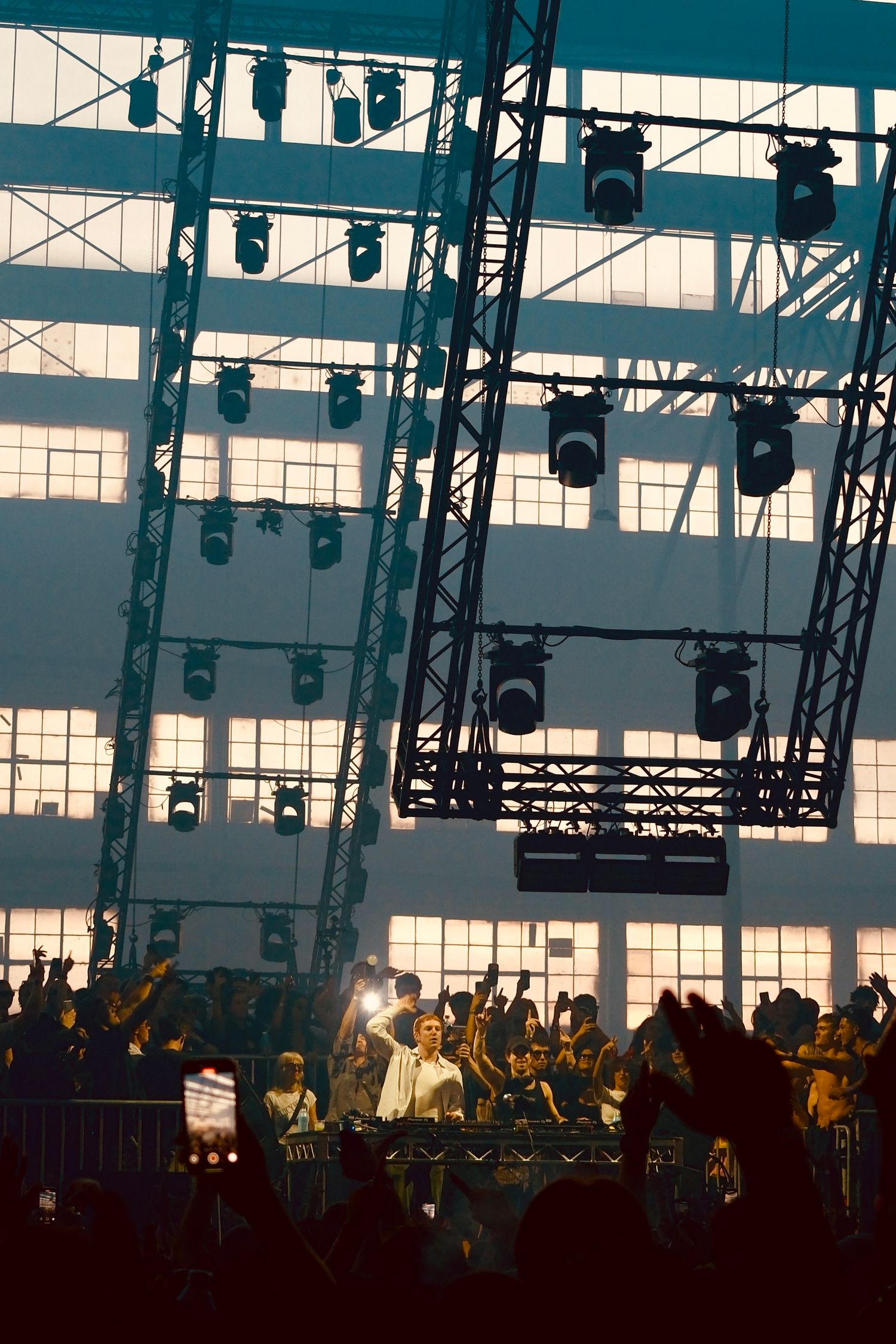 DJ performs on stage at an indoor warehouse concert with rigging and spotlights overhead.