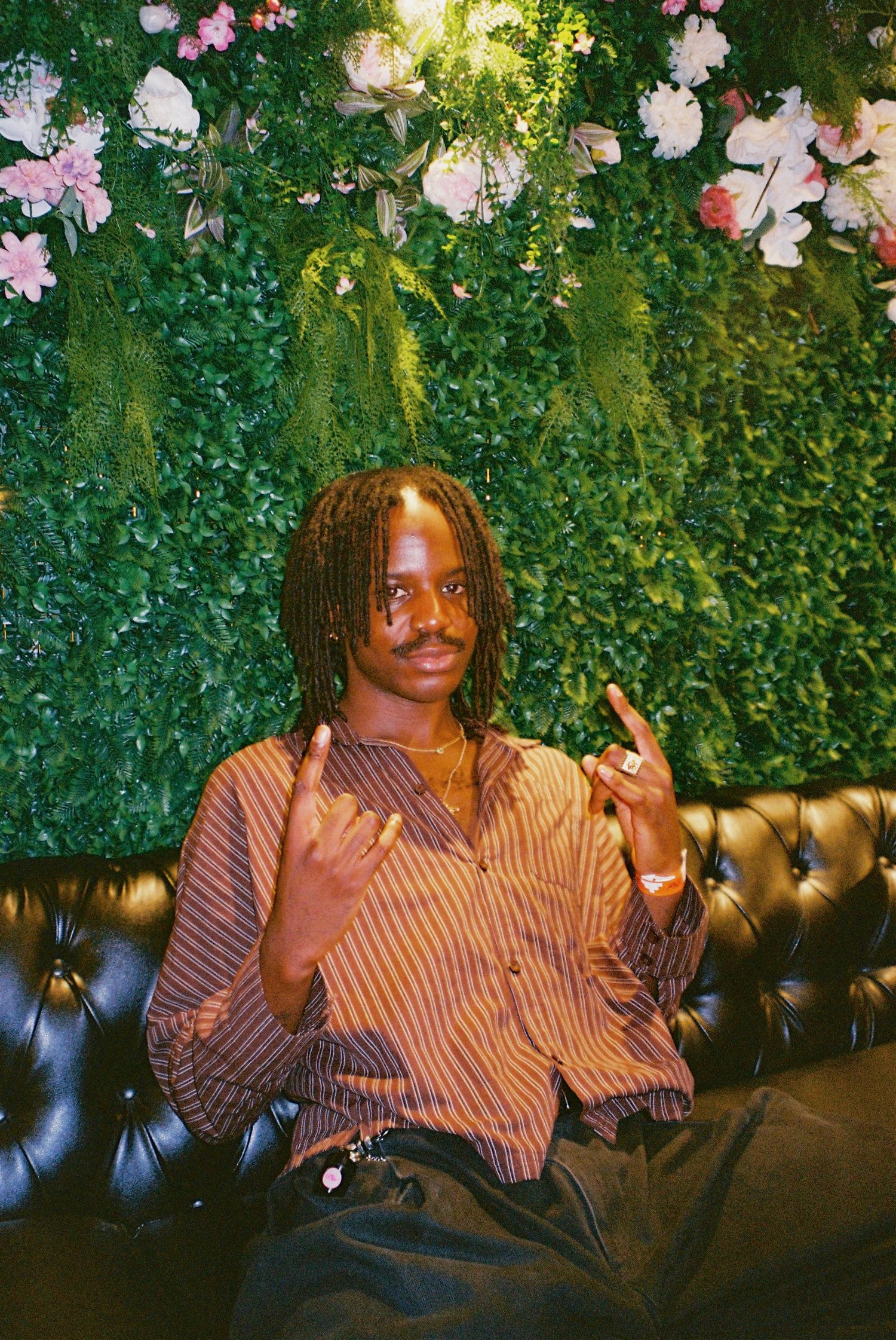 Young man in striped shirt sitting on leather sofa with floral wall backdrop.