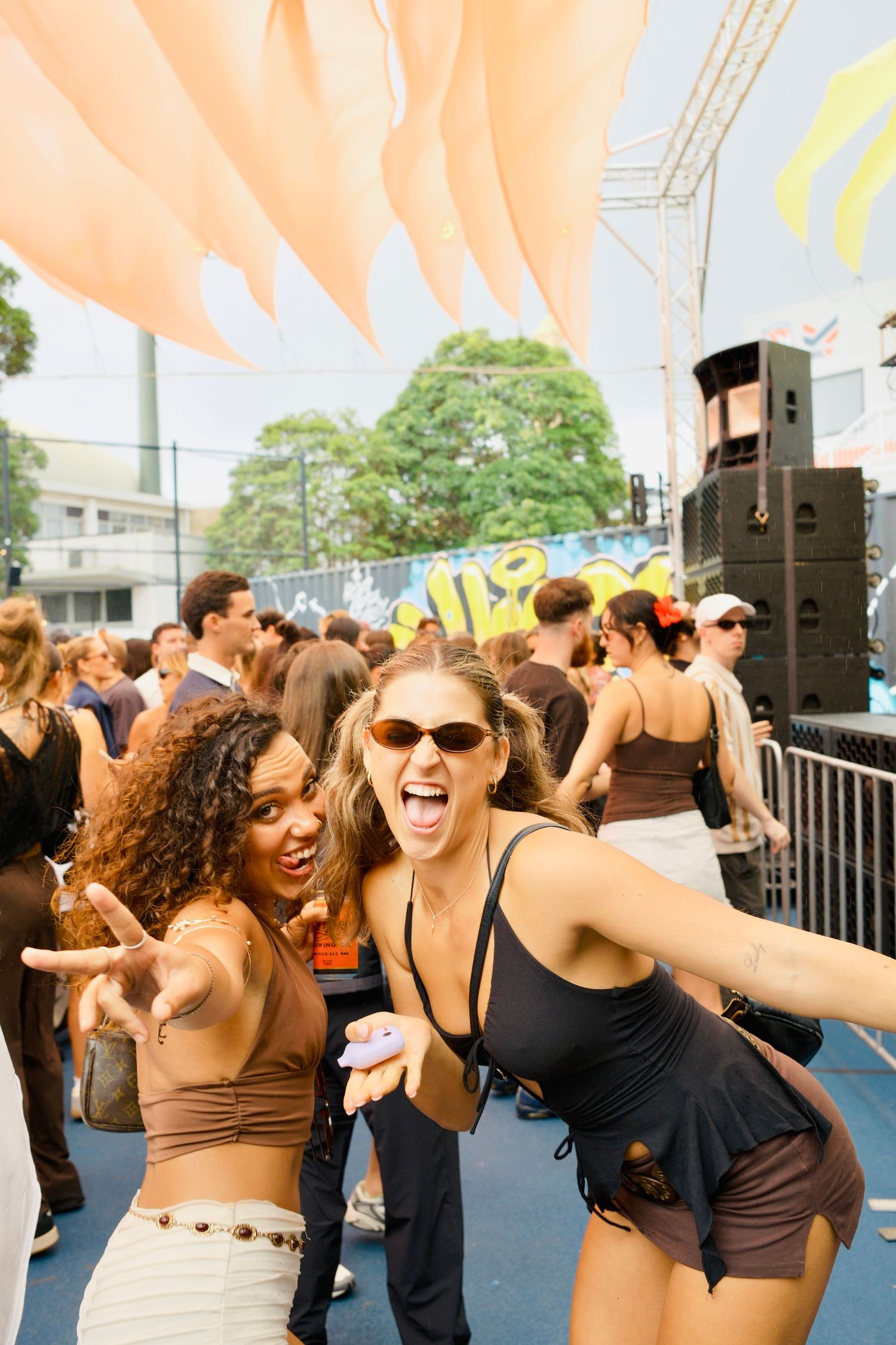Two excited young women posing and making peace signs at a vibrant outdoor music festival.