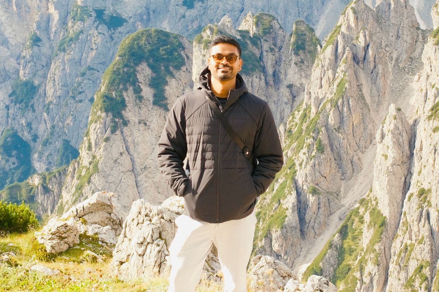 Man in black puffer jacket standing confidently on a rocky Alpine mountain trail with cliffs behind.