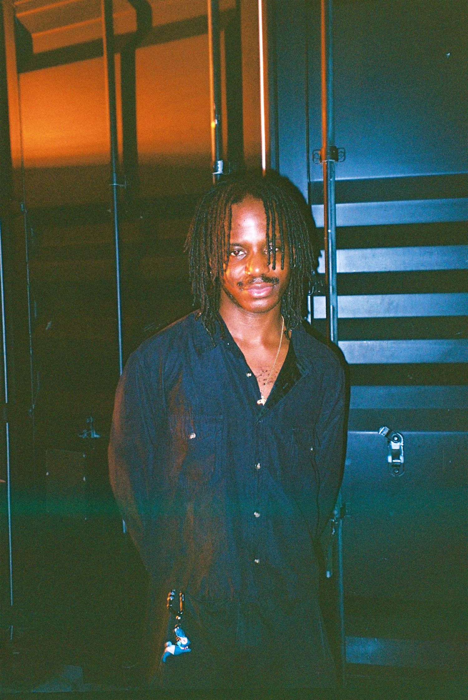Young man with dreadlocks in dark shirt posing indoors near staircase in dim lighting.