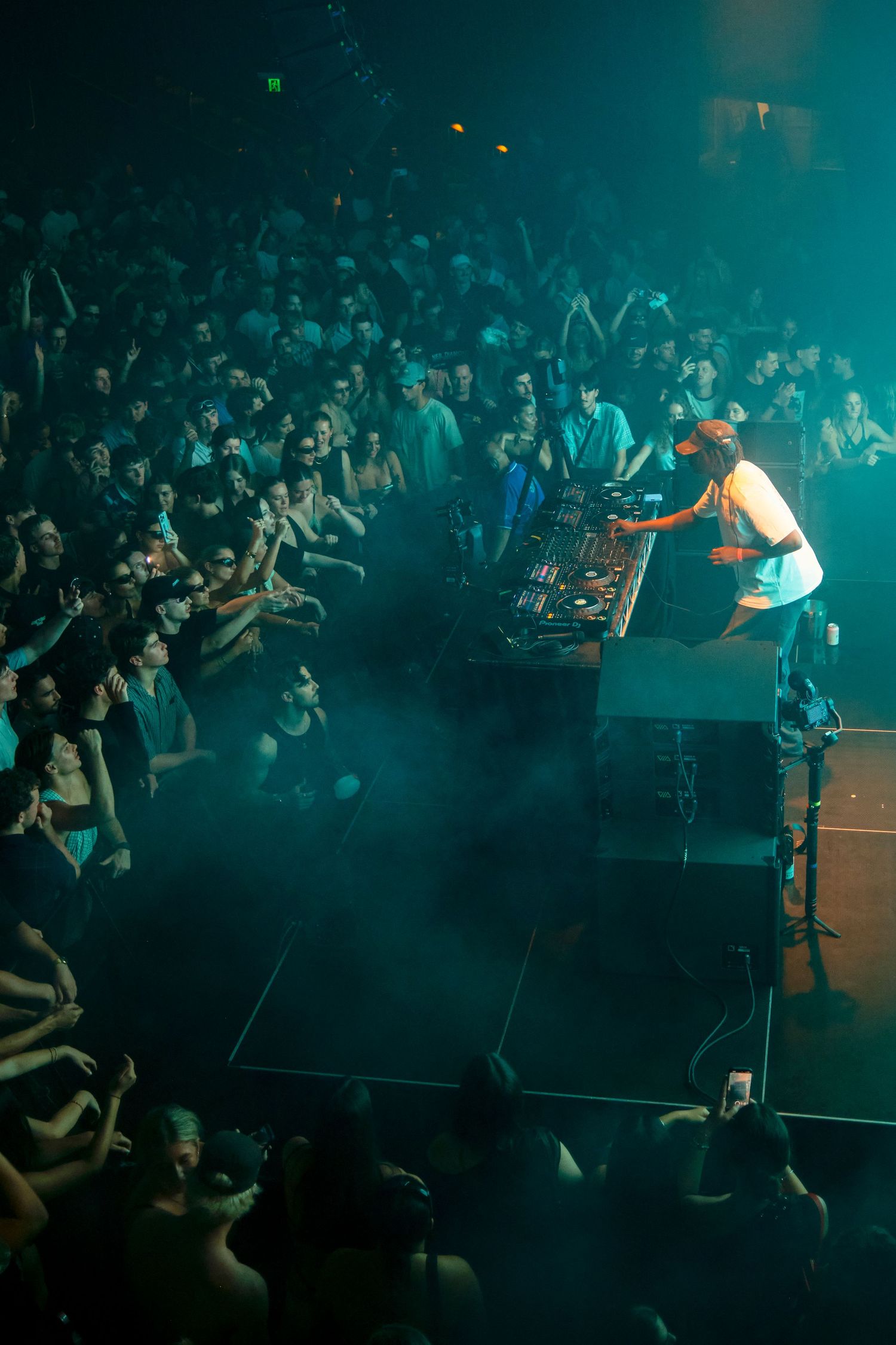 DJ performing on stage with turntables surrounded by a large crowd in a dark, fog-filled concert venue.