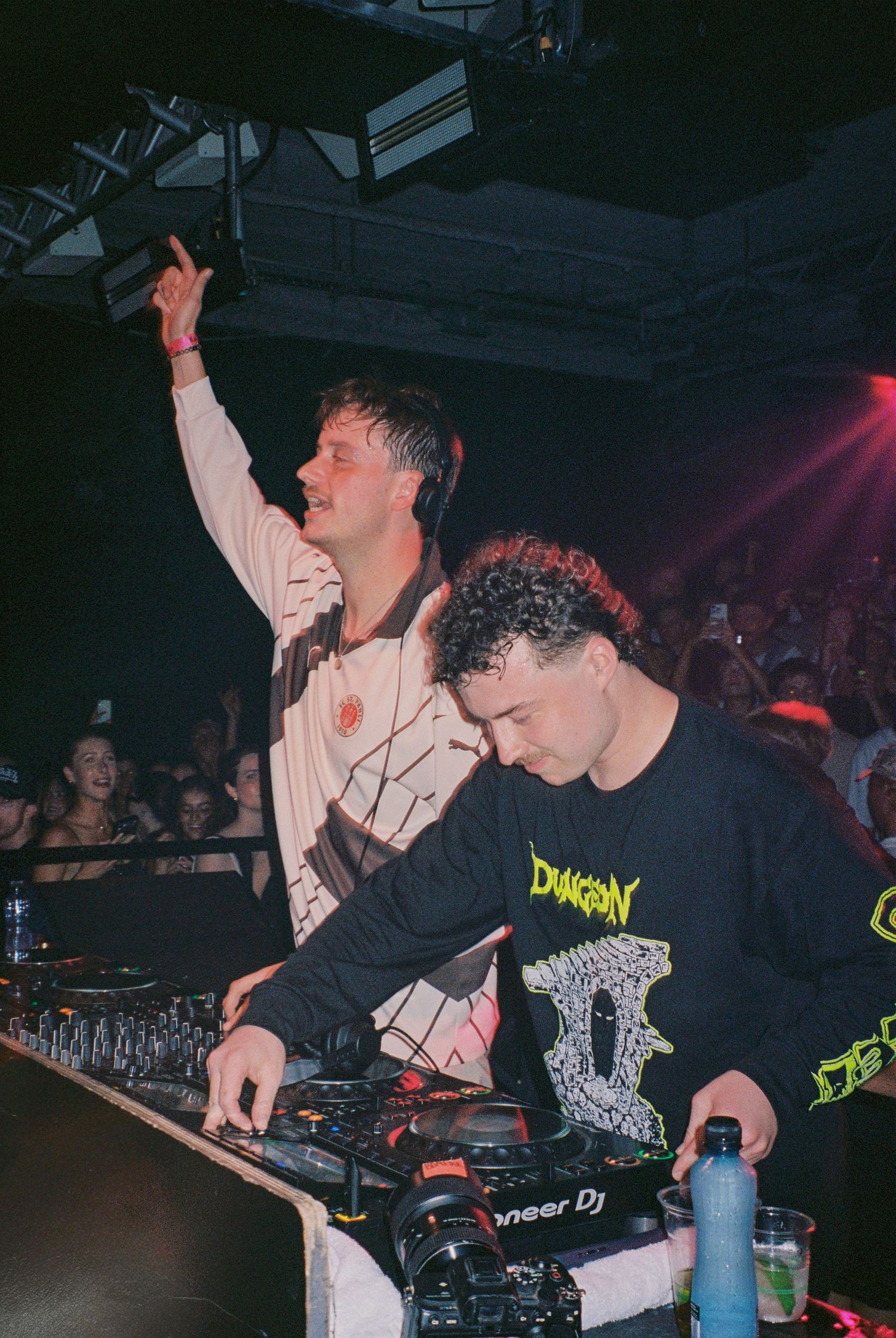 Two DJs perform at a nightclub using Pioneer DJ equipment while an energetic crowd watches in the background.