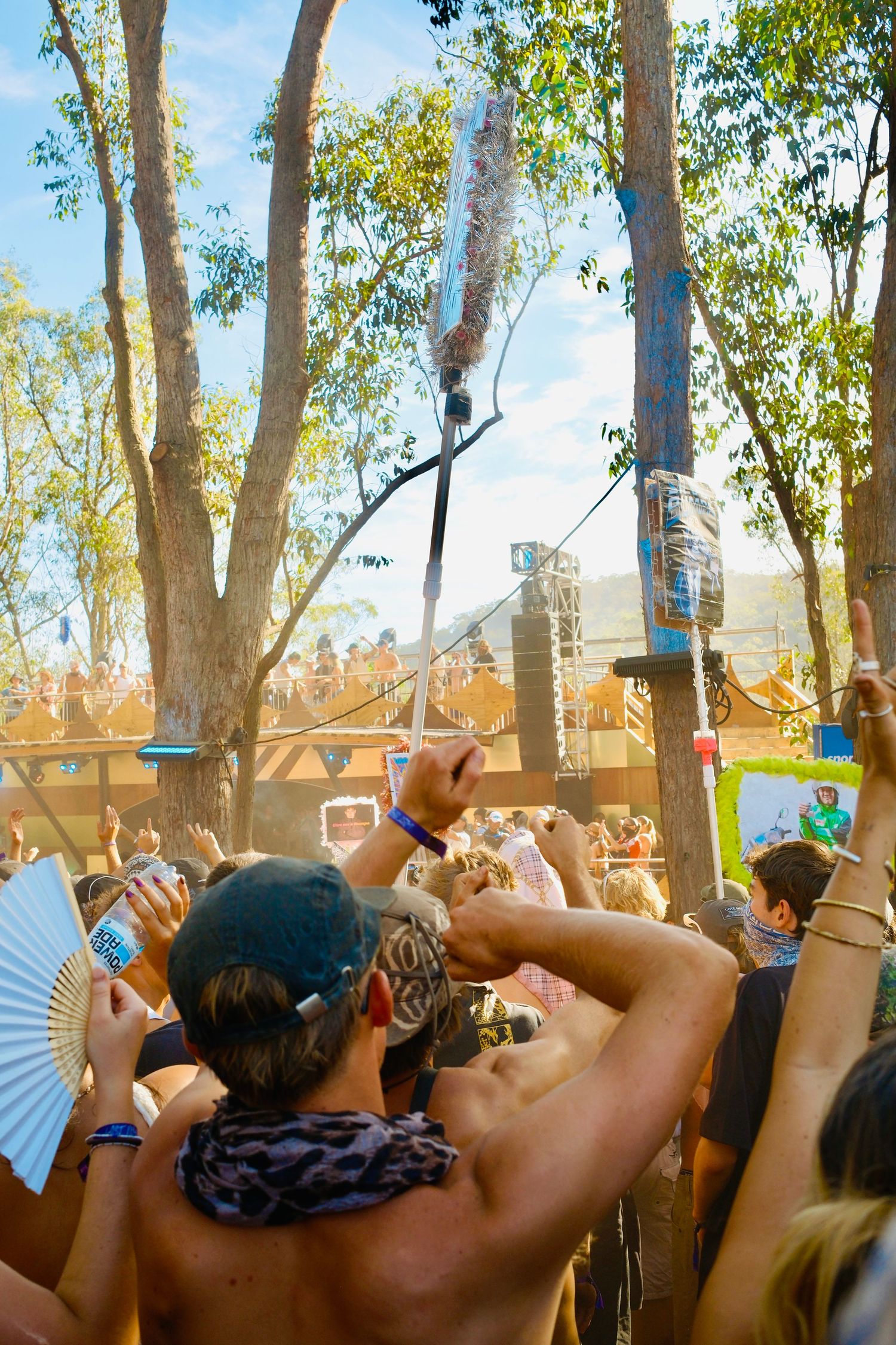 Festival crowd dancing outdoors beneath eucalyptus trees with water misting on a sunny day.