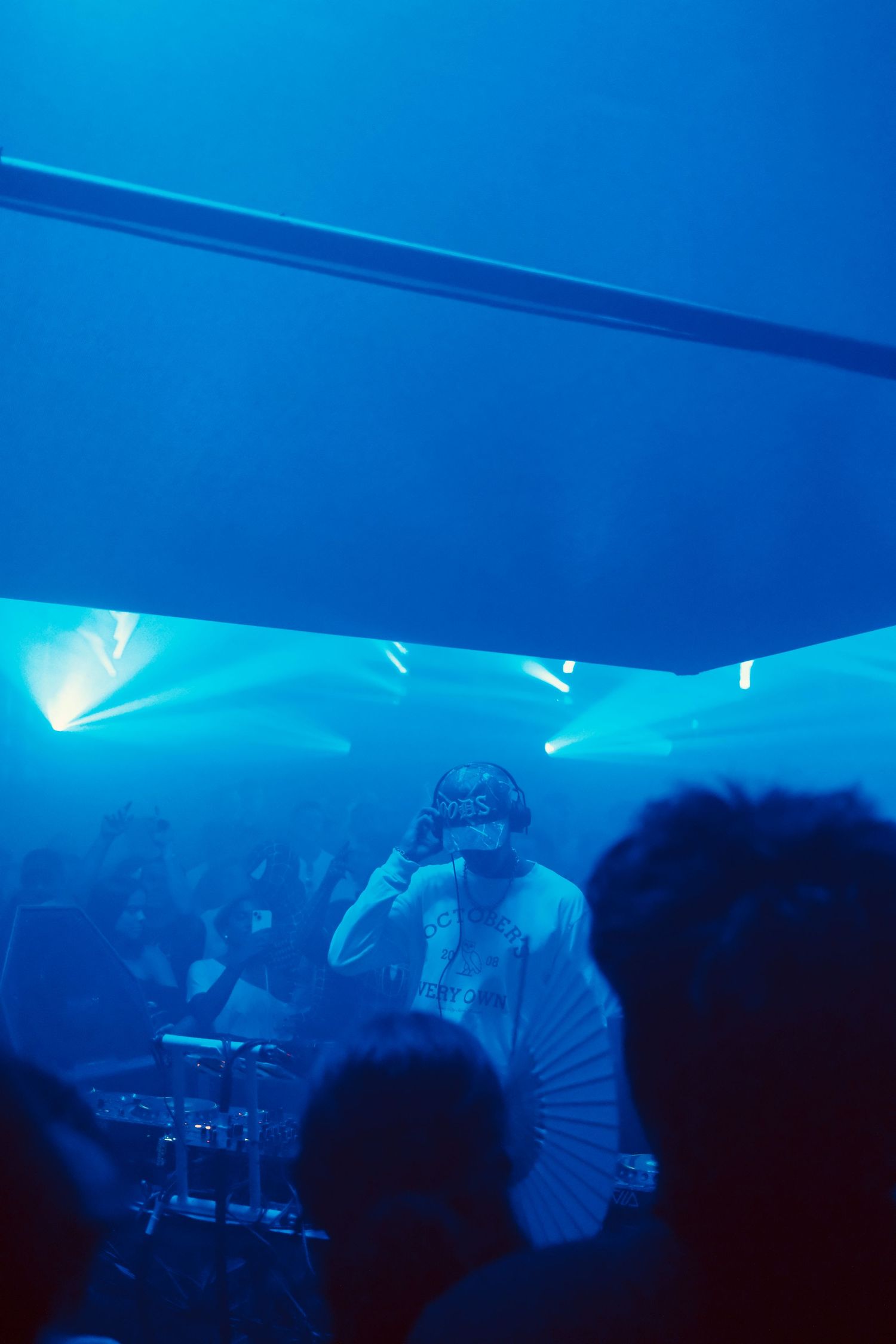 DJ in white outfit and cap performing at decks surrounded by blue fog in a nightclub.