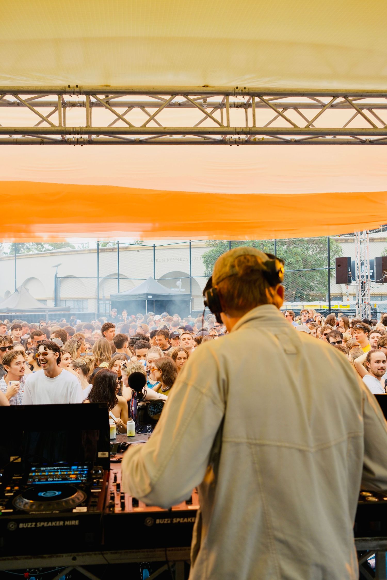DJ performing on stage facing a large crowd at an outdoor music festival under an orange canopy.