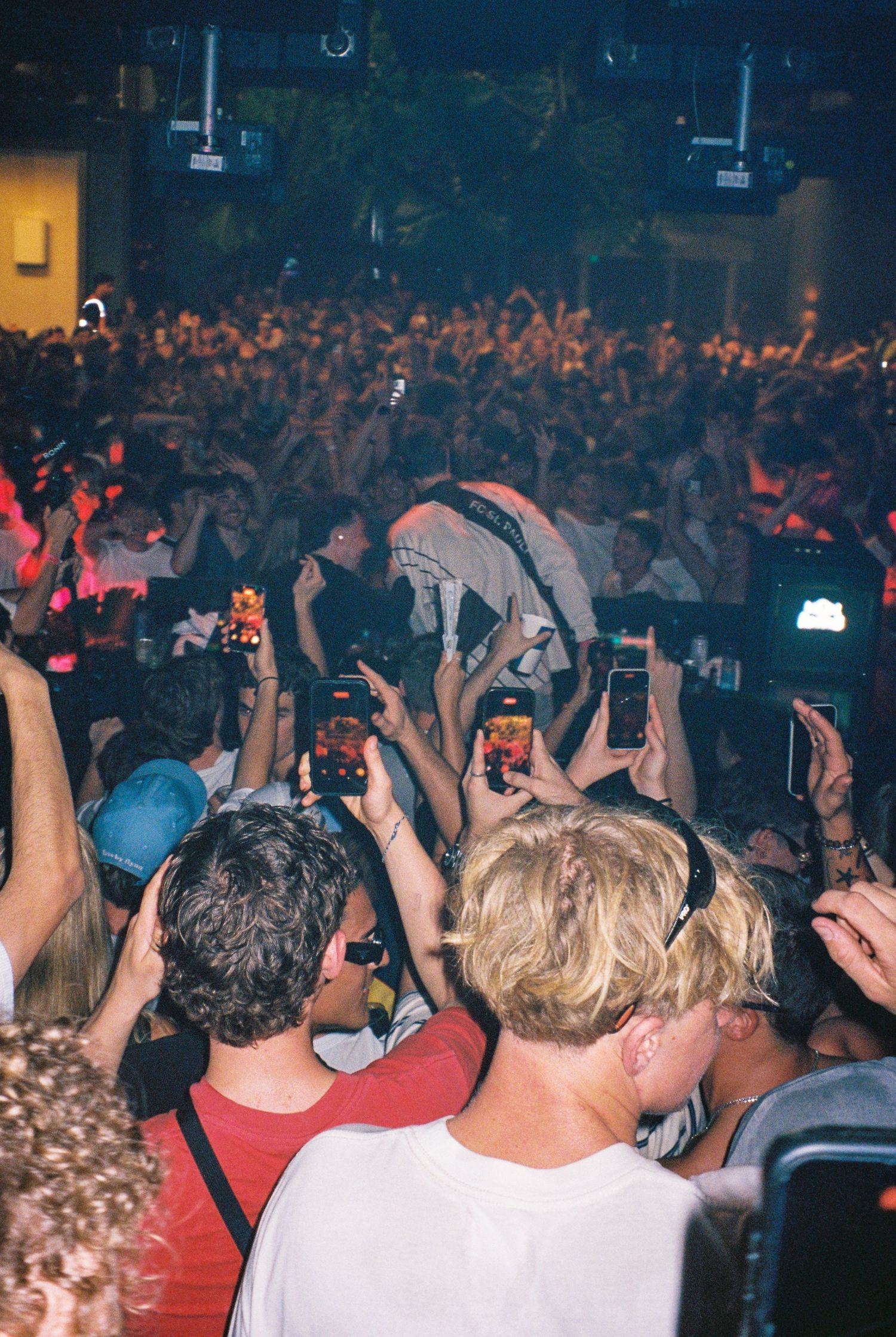 Crowd surfing at a packed indoor concert with fans filming on smartphones under bright stage lights.