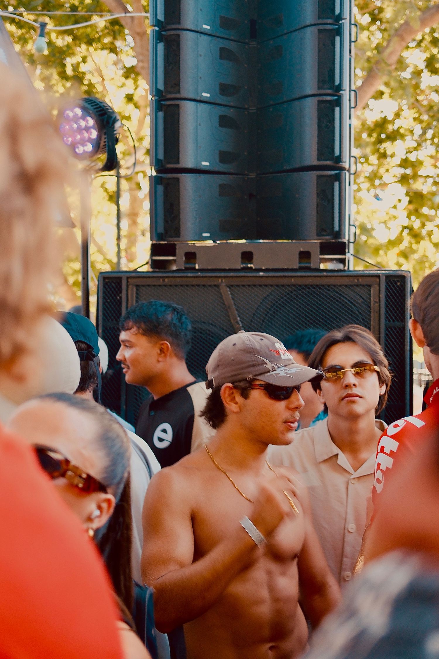 Shirtless young man stands near large speaker stacks at a sunny outdoor music festival.