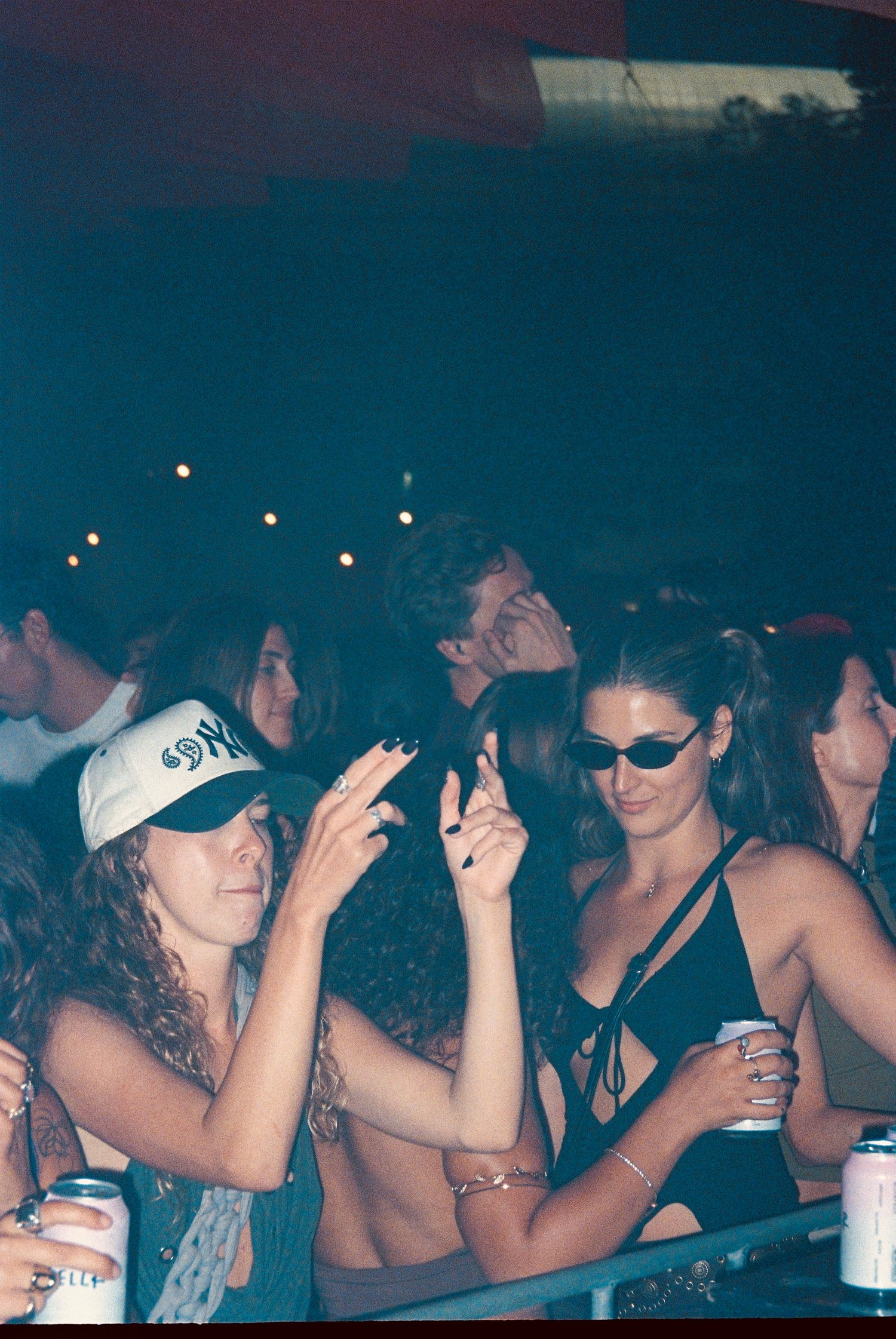 Festival crowd at night with two women making hand gestures holding drinks near barrier.