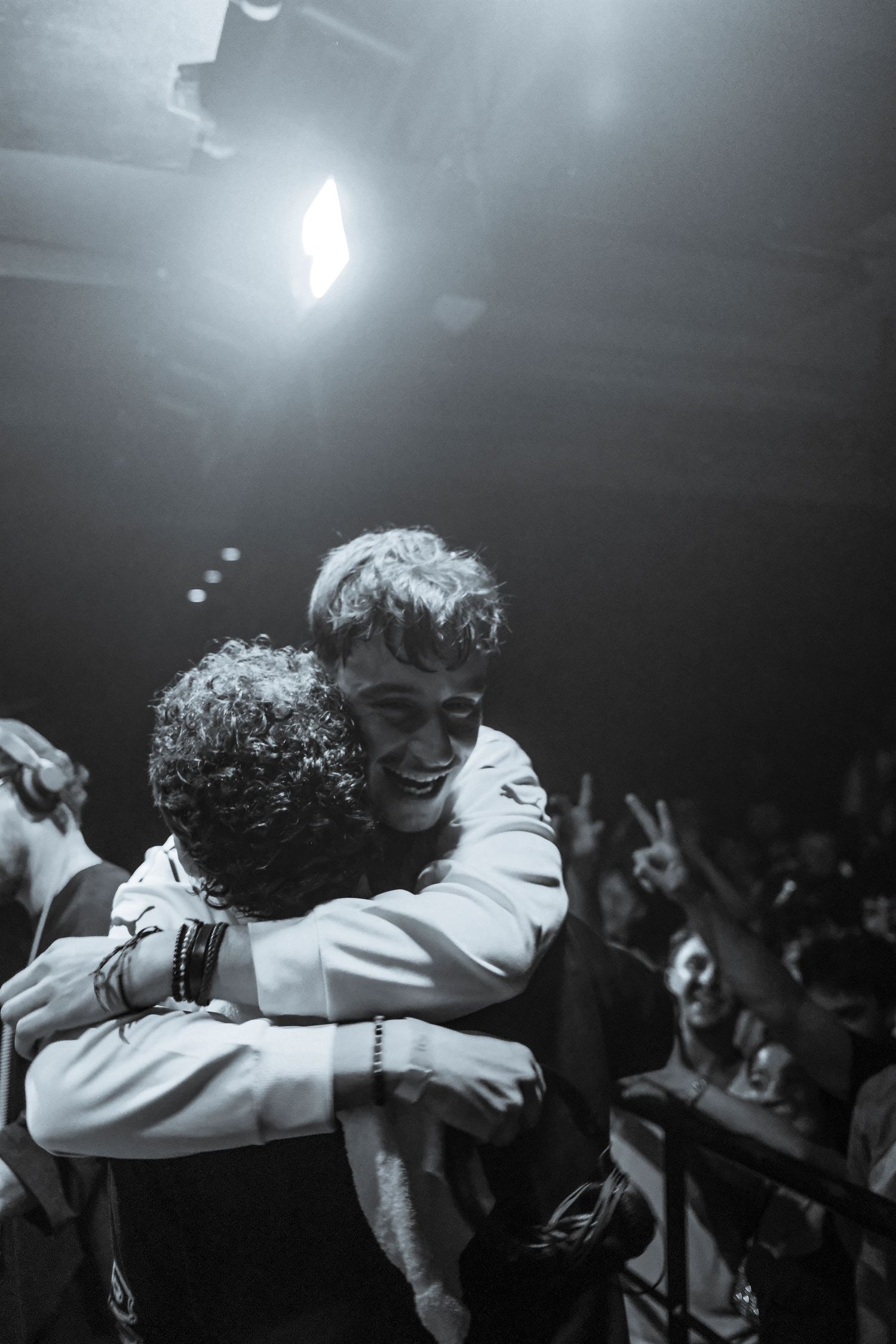 Two people share a warm hug on stage at a concert, smiling joyfully in black and white photography.