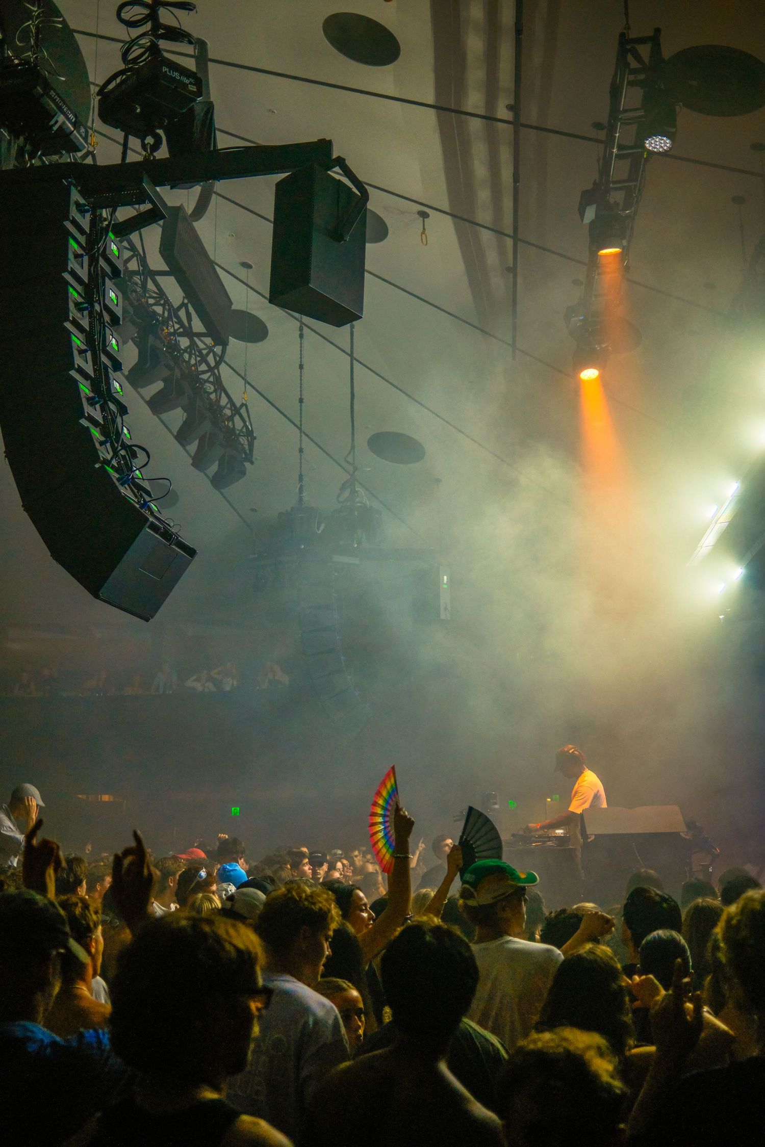 Crowd enjoys a live concert with dramatic stage lighting, fog effects, and a DJ performing on stage.