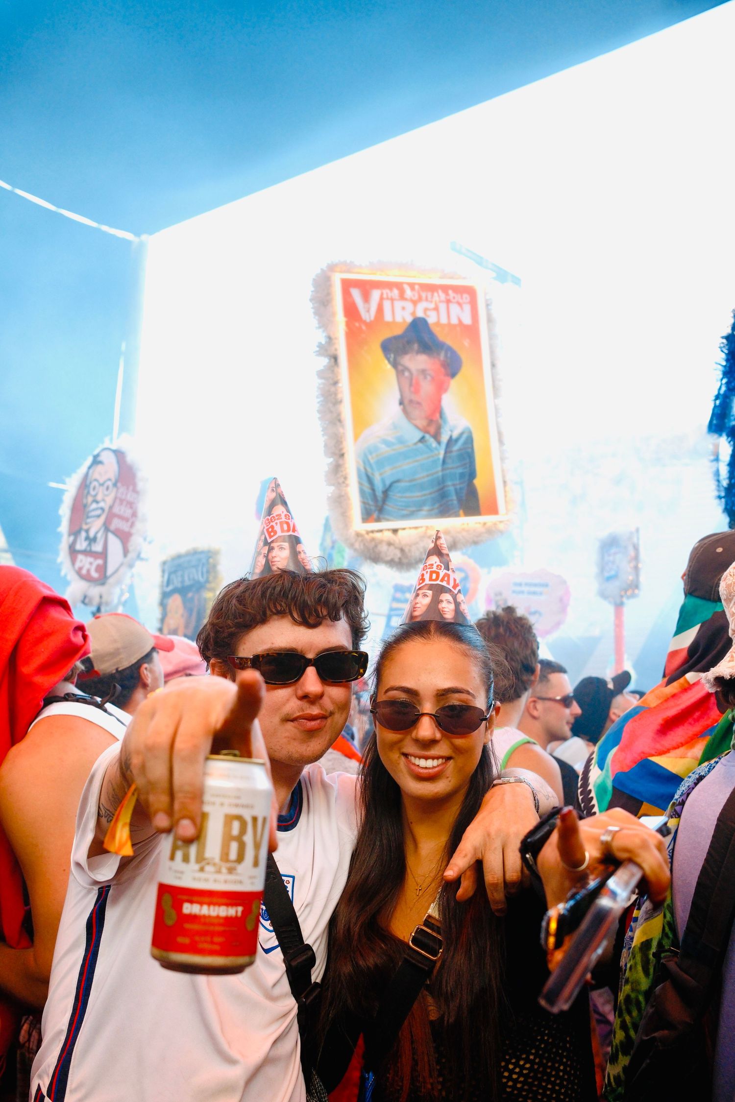 Couple pointing at camera holding drinks at a lively outdoor music festival.
