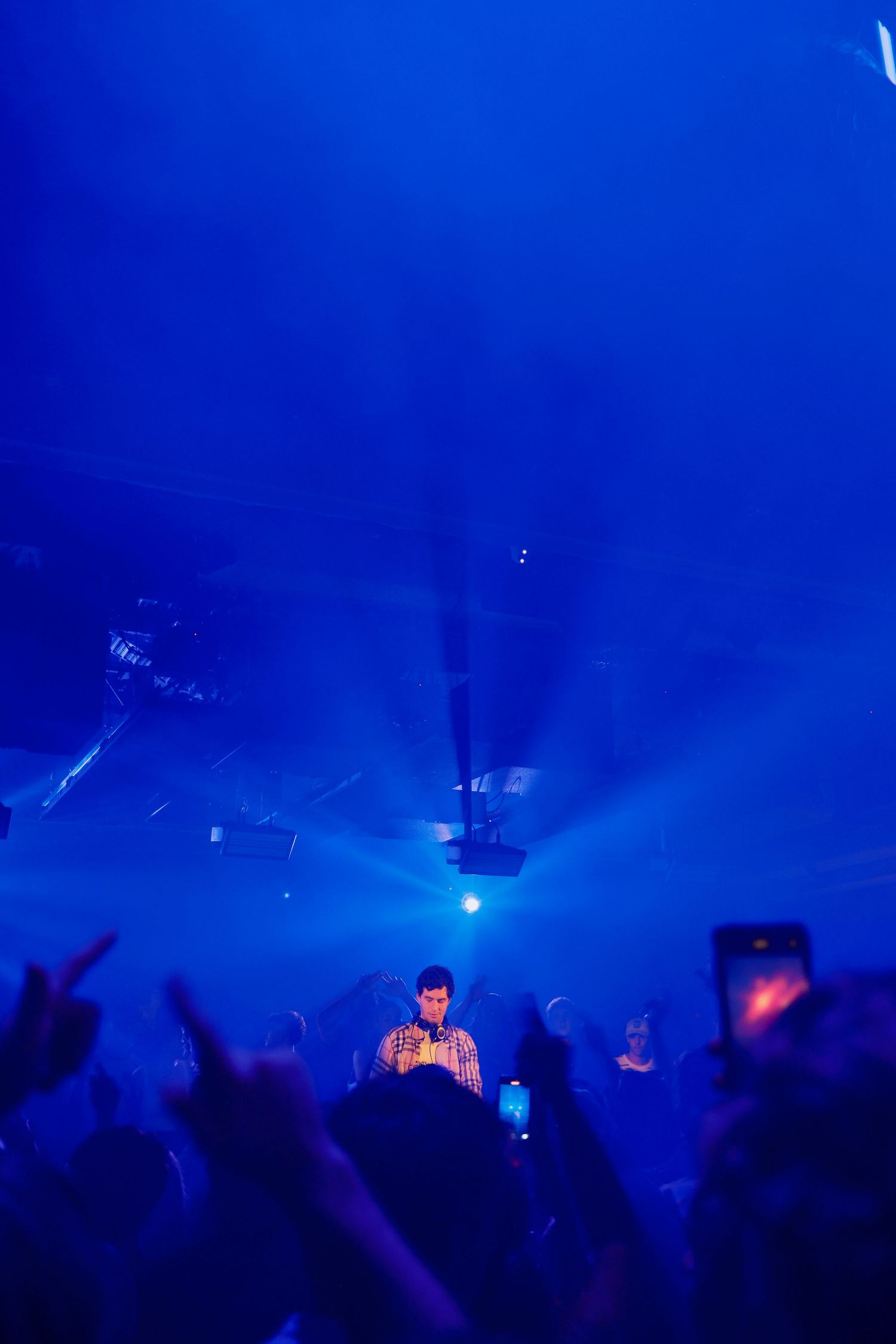 DJ performing on stage as crowd raises hands under vivid blue light beams in a packed nightclub.