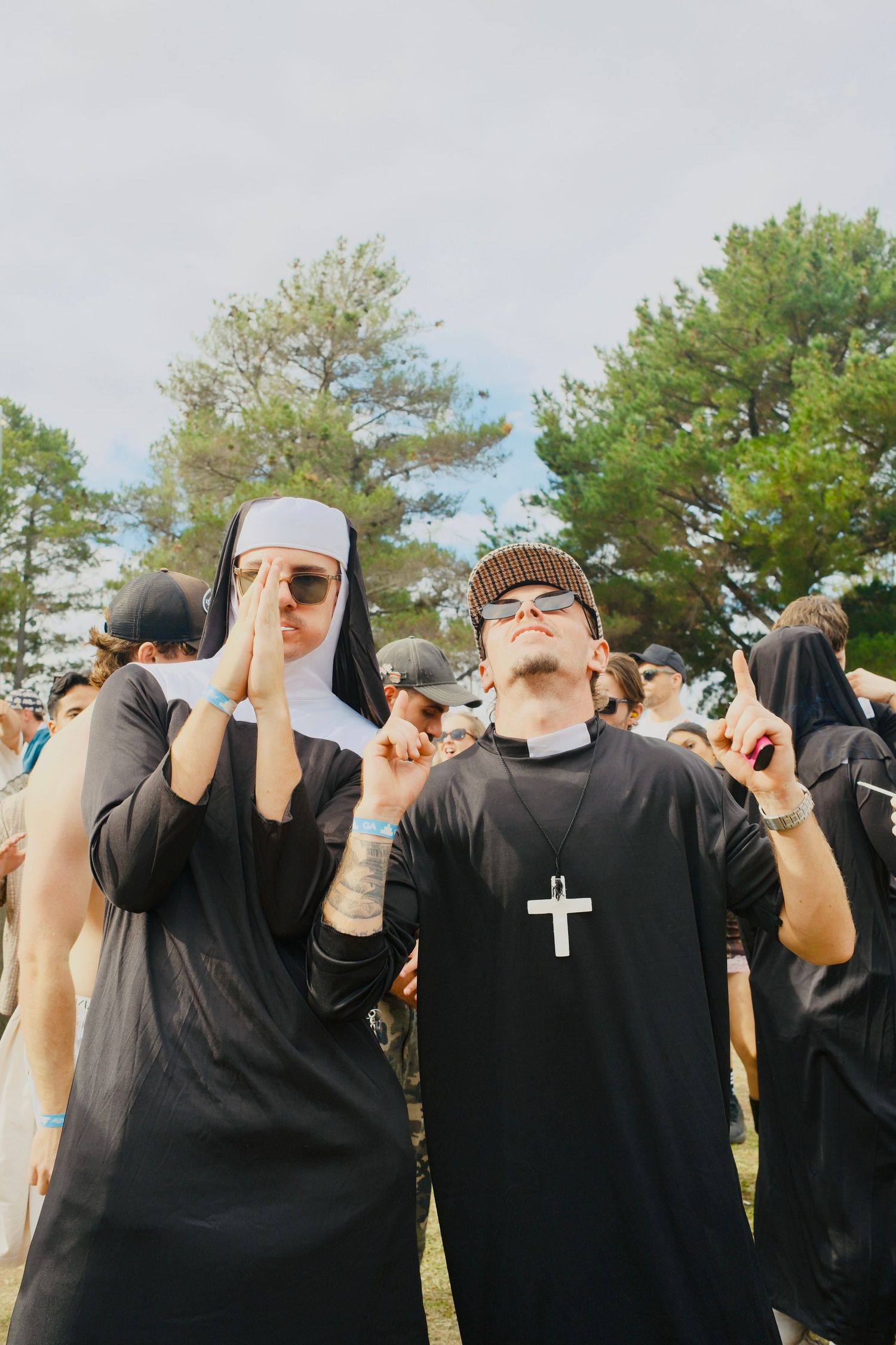 Two people dressed in nun and priest costumes posing together at an outdoor music festival crowd.