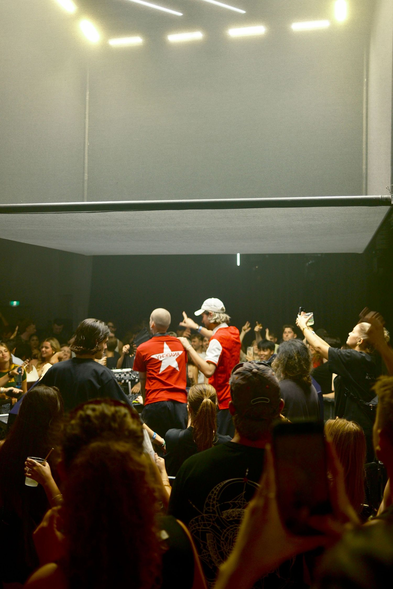 DJ performing for a packed indoor crowd at a nightclub with overhead lighting and raised hands.