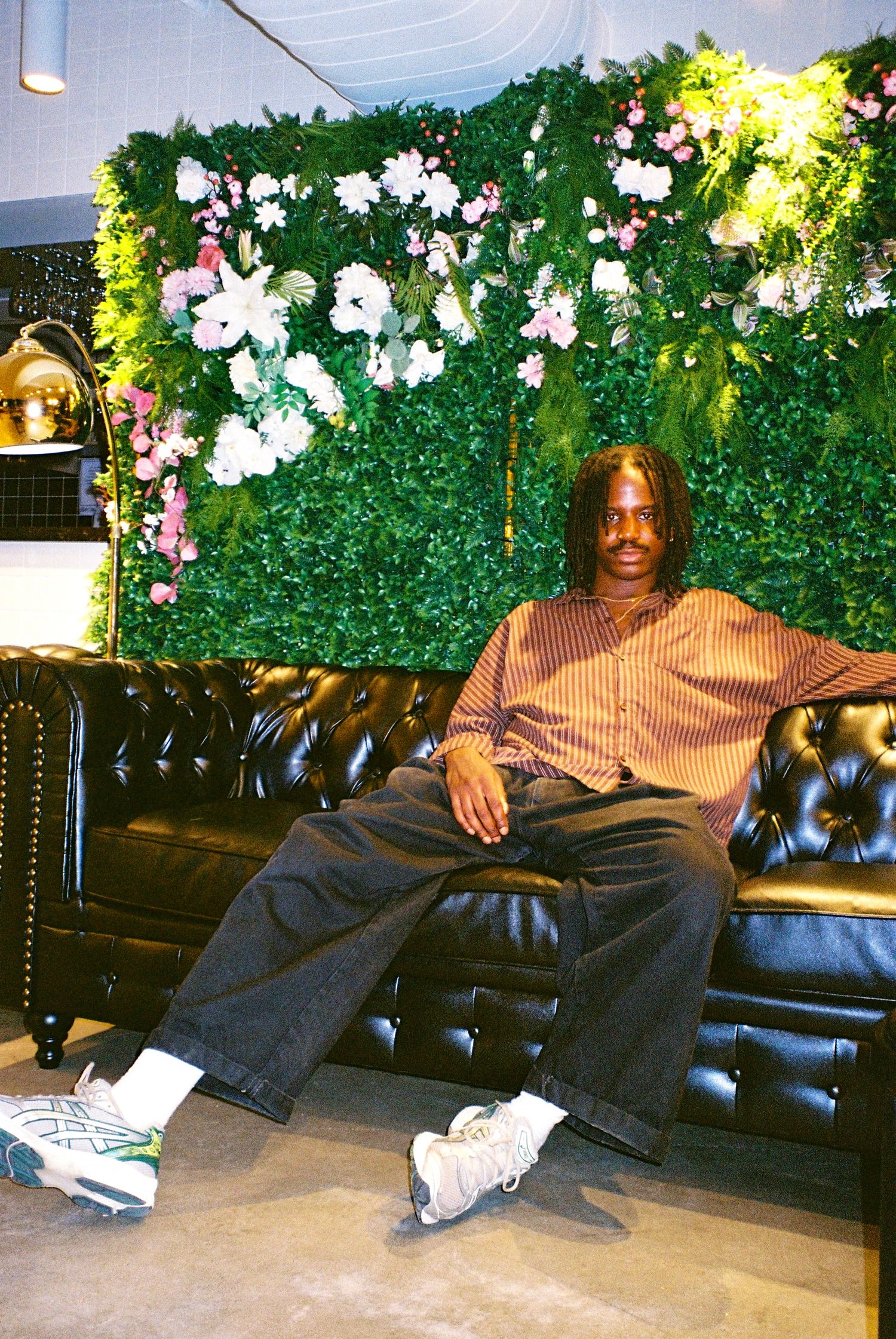 Man in striped shirt and dark jeans relaxing on black leather sofa with green floral wall.
