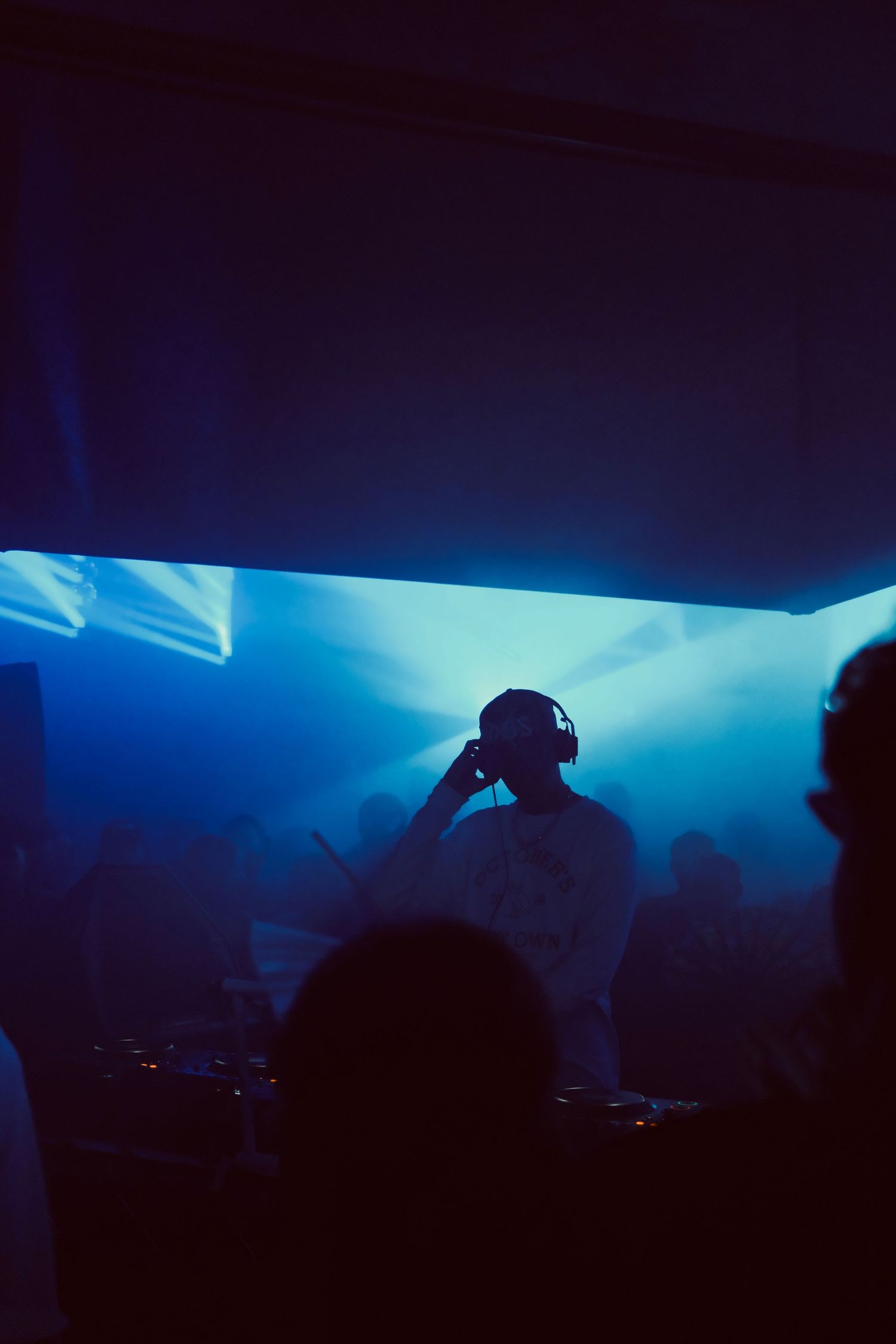 DJ silhouette wearing headphones performing at a nightclub with blue haze and spotlight behind.