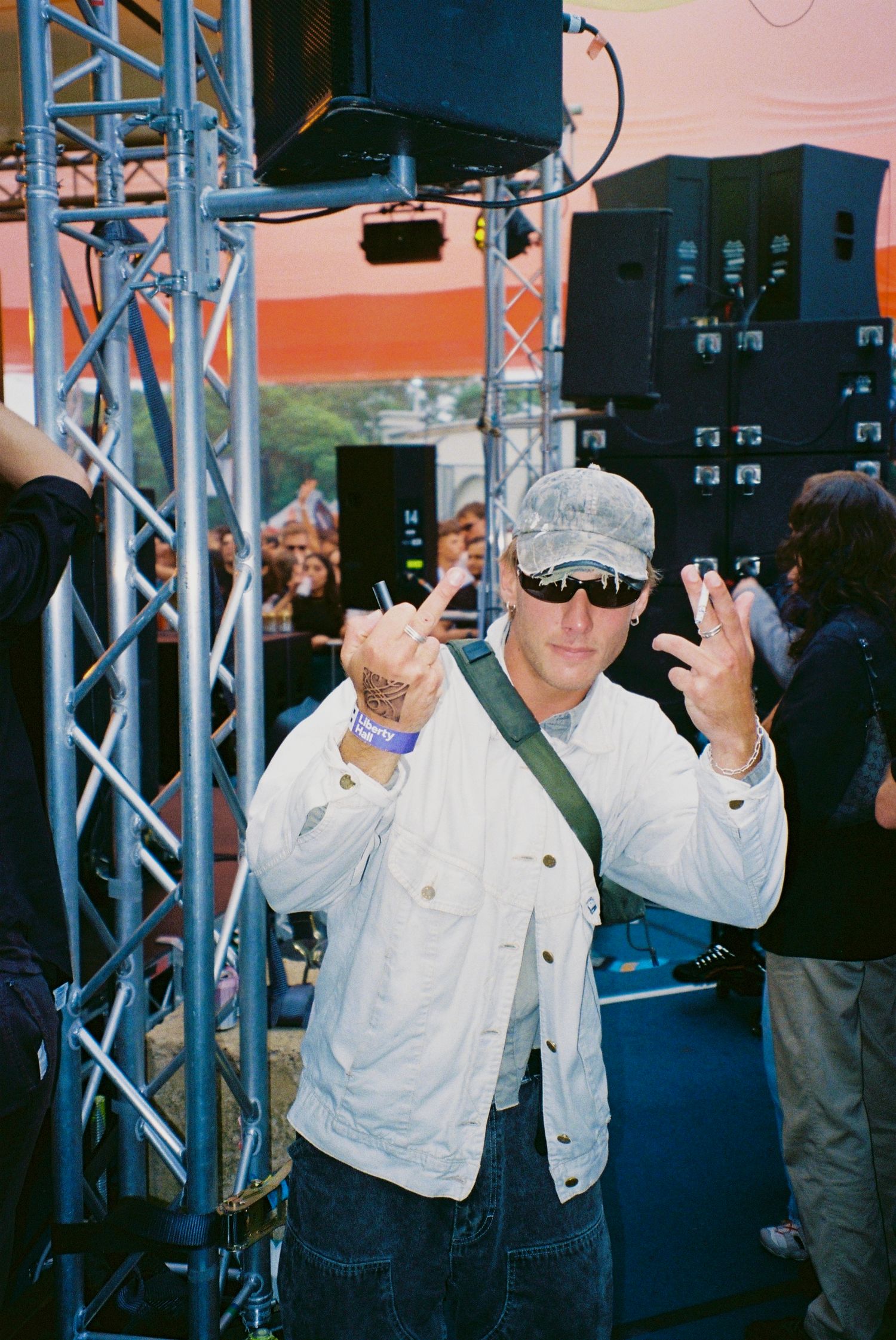 Man in white denim jacket and camo cap making hand gestures near festival stage speakers.