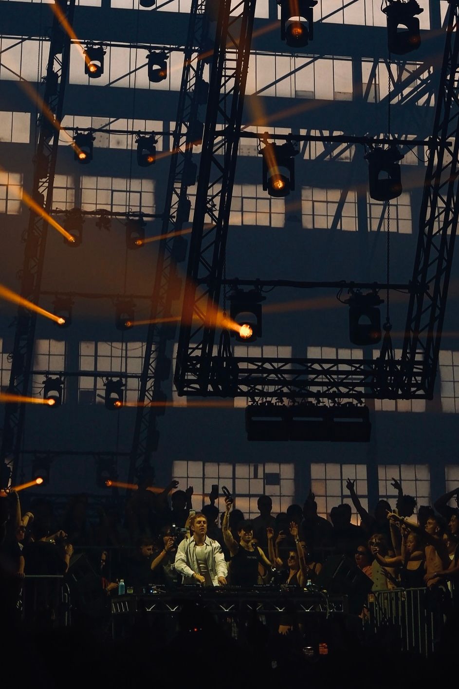 DJ performing under golden stage lights in a large industrial warehouse concert venue.