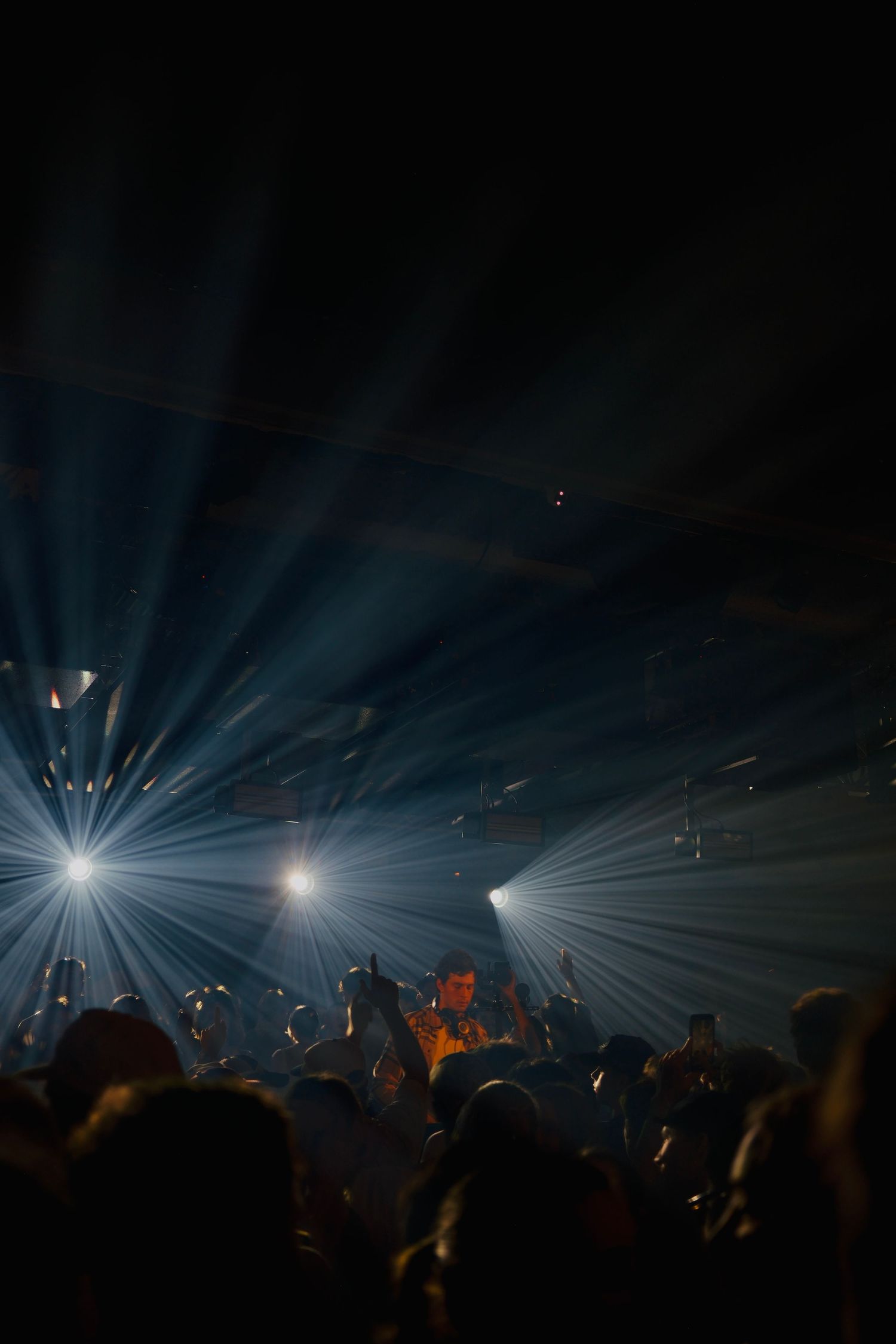 Bright spotlight beams pierce through darkness above a packed nightclub crowd at a live event.
