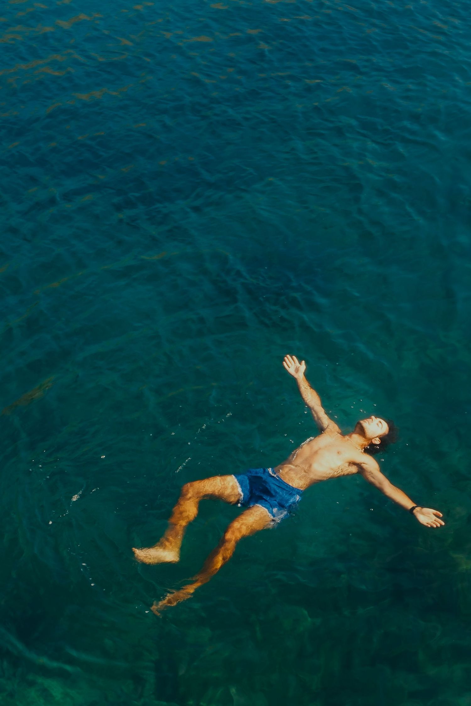 Aerial view of a man floating on his back in clear turquoise Mediterranean sea water wearing blue swim shorts.