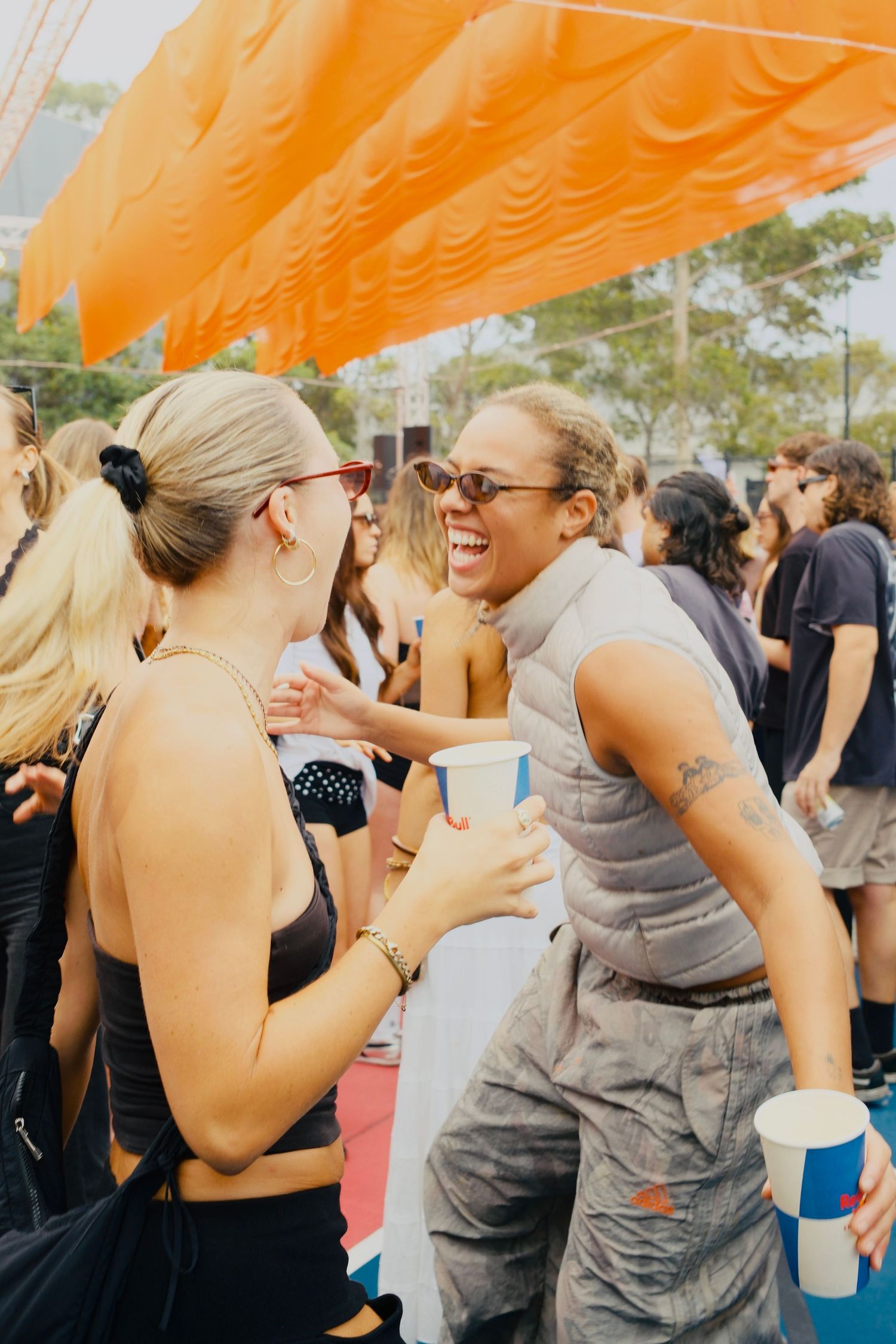Two women laughing and socializing with drinks under an orange canopy at an outdoor festival.