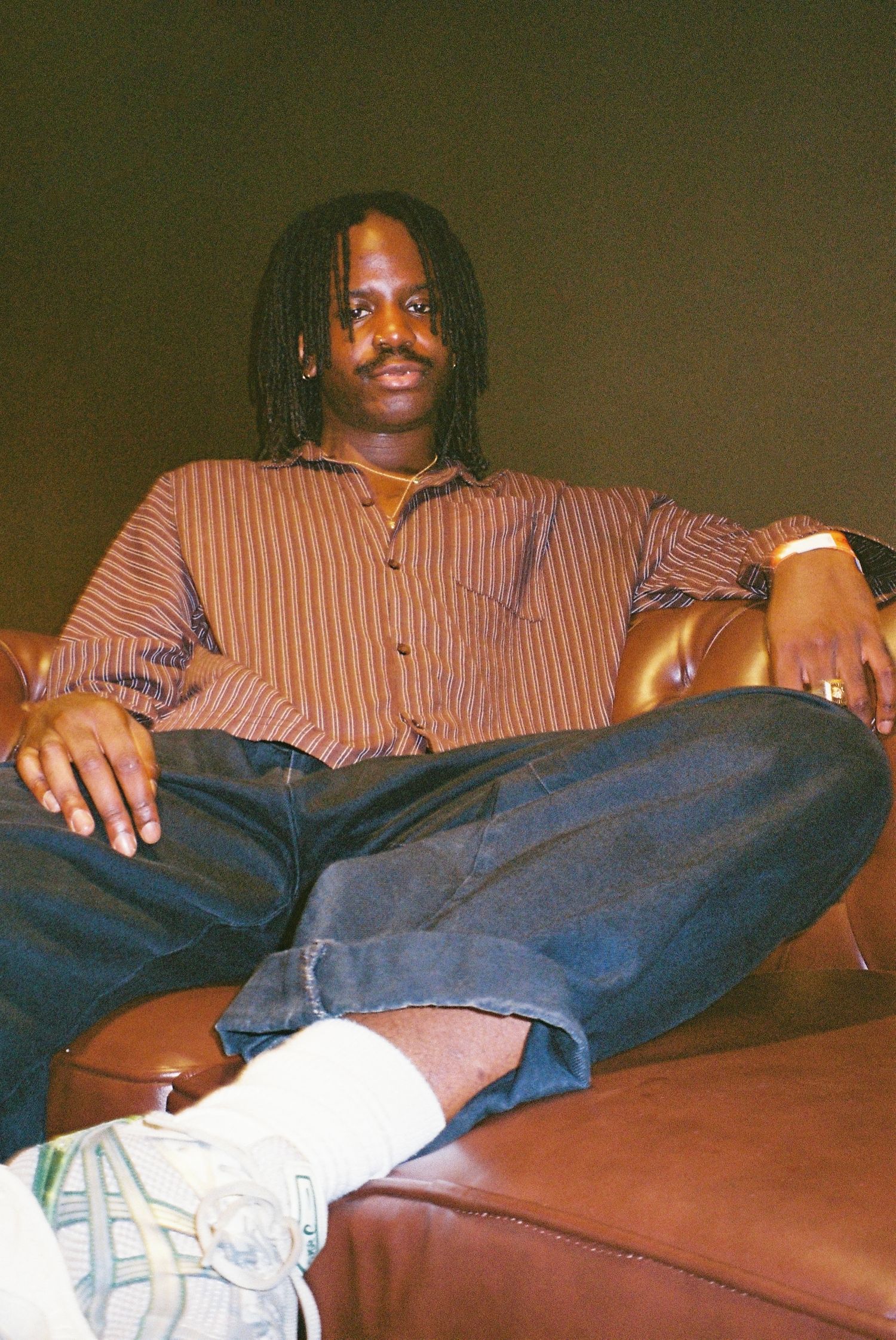 Man with dreadlocks in striped shirt sitting cross-legged on brown leather sofa.