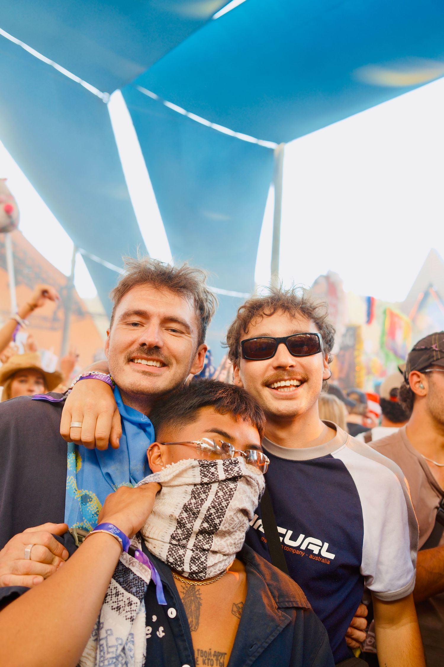 Three friends smiling and posing together at an outdoor music festival under a blue tent canopy.