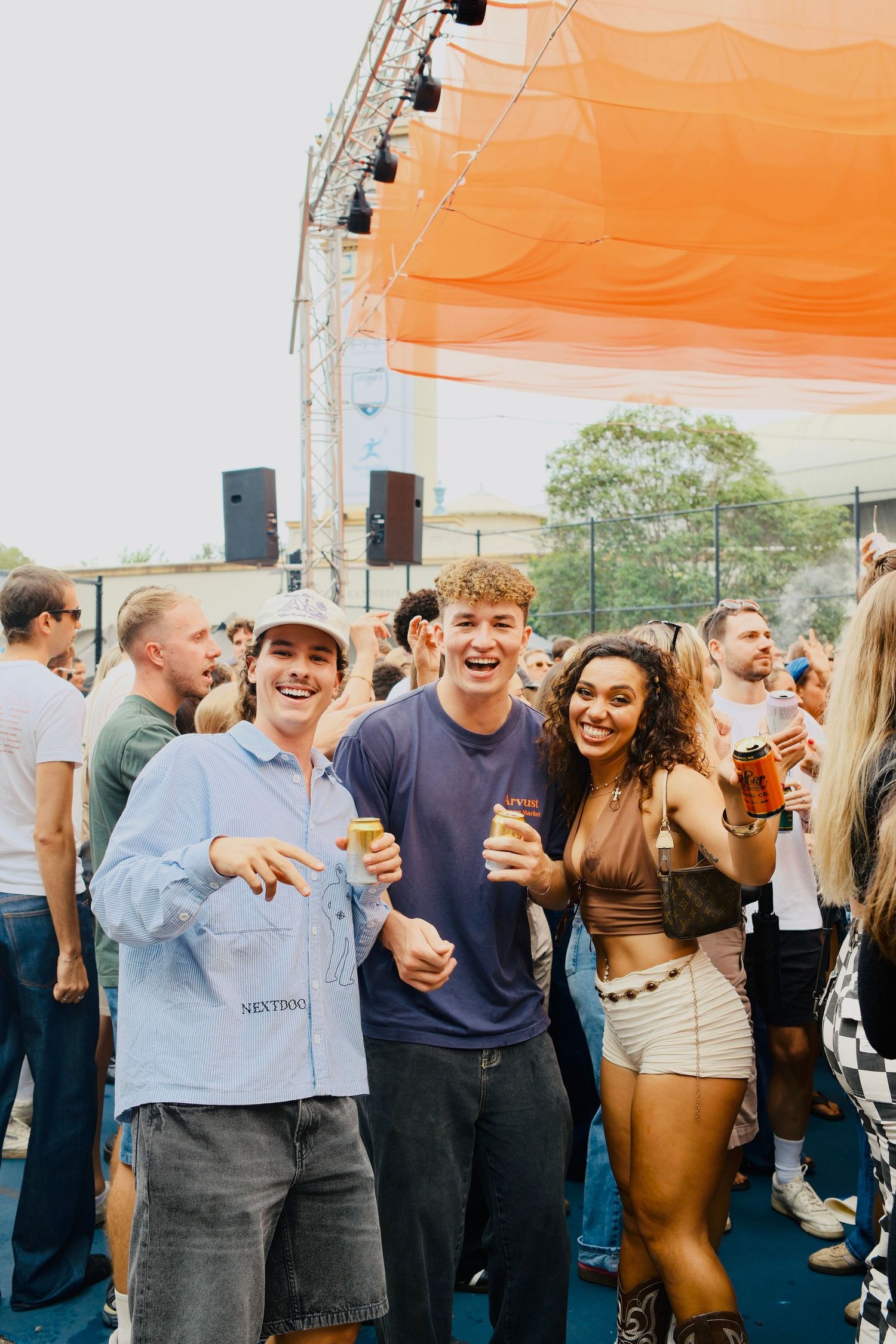 Three smiling friends posing together at a lively outdoor music festival with a crowd behind them.