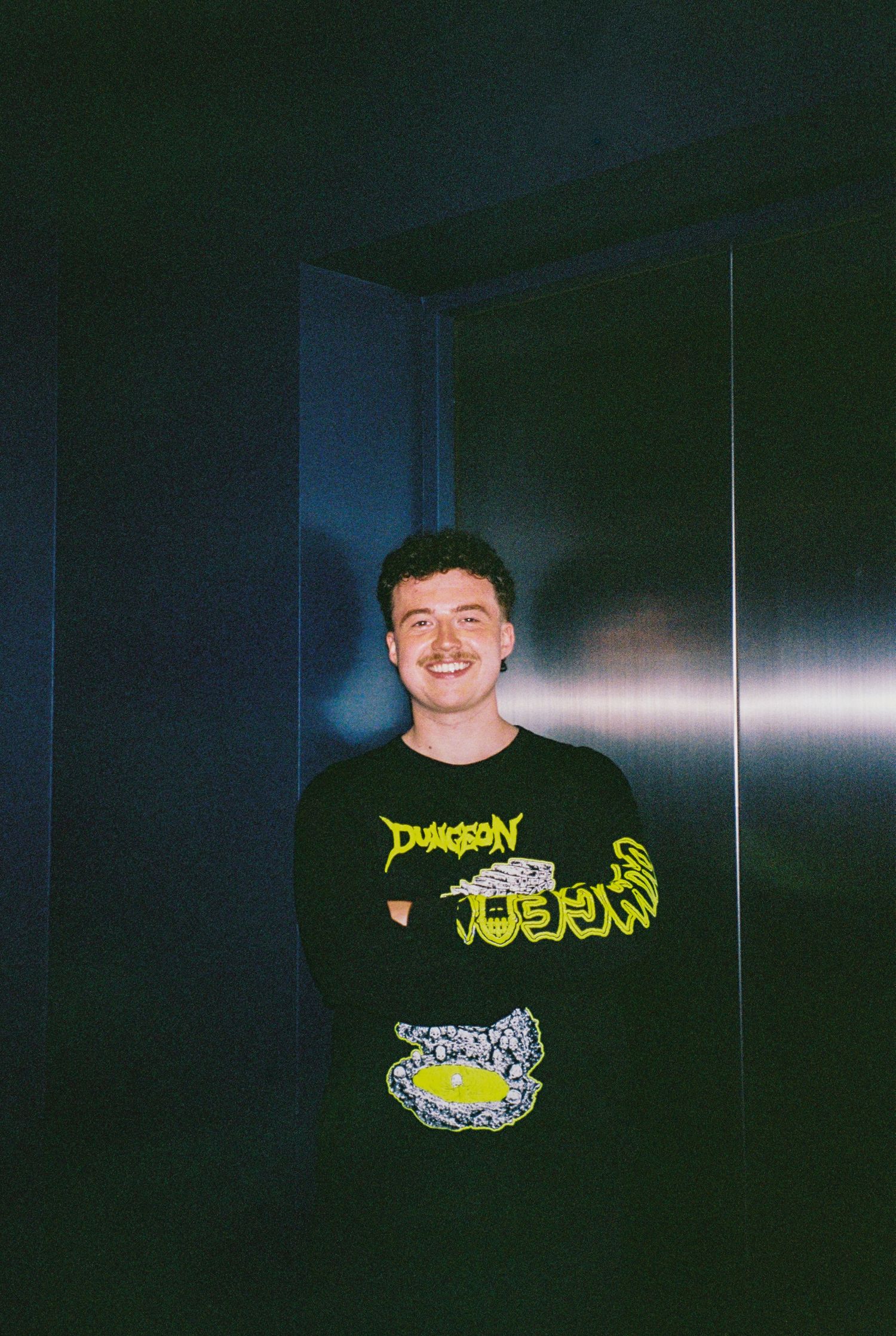 Smiling young man in black Dungeon long-sleeve shirt standing in a dimly lit doorway.