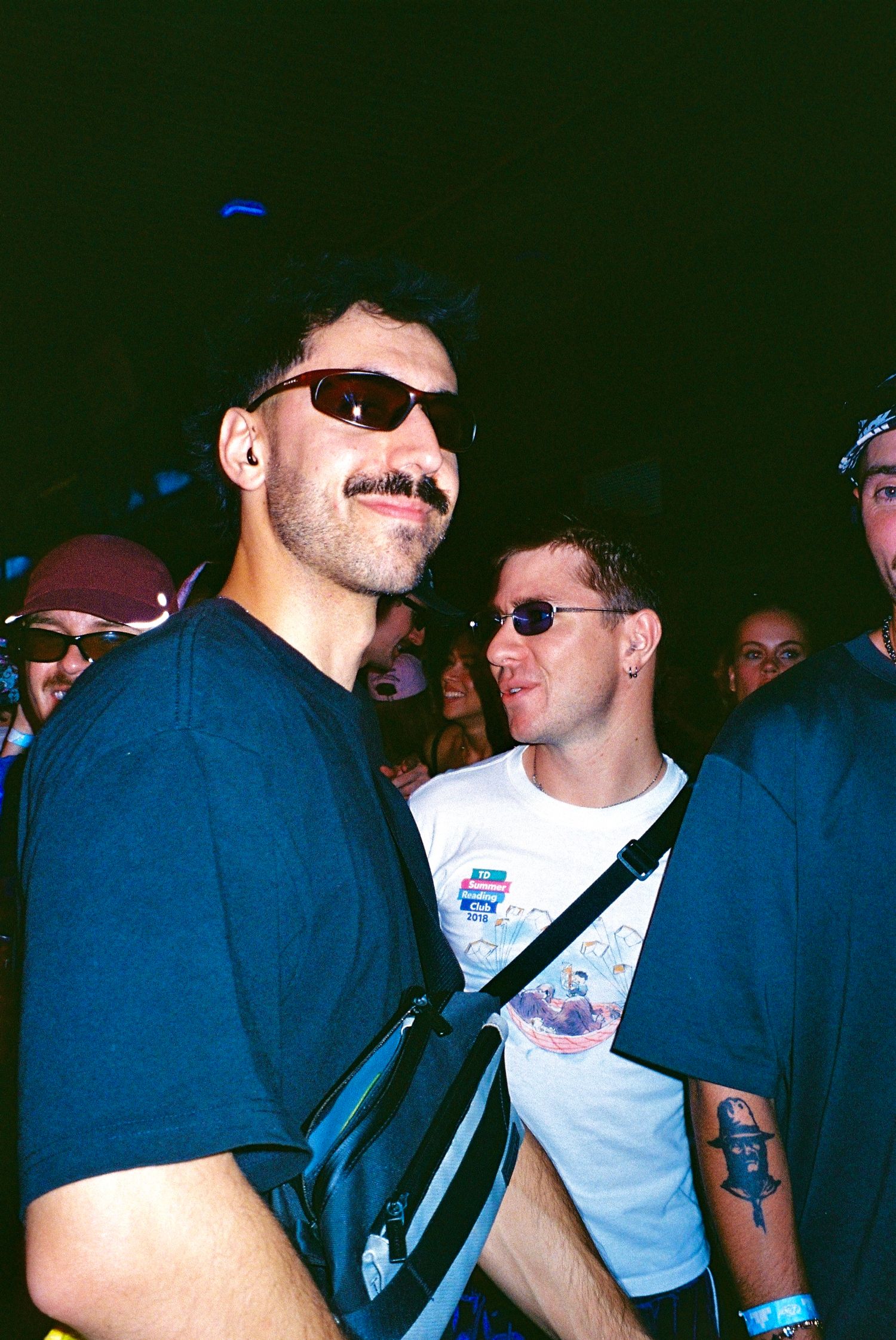 Tall man with mustache wearing wraparound sunglasses and crossbody bag at music event.
