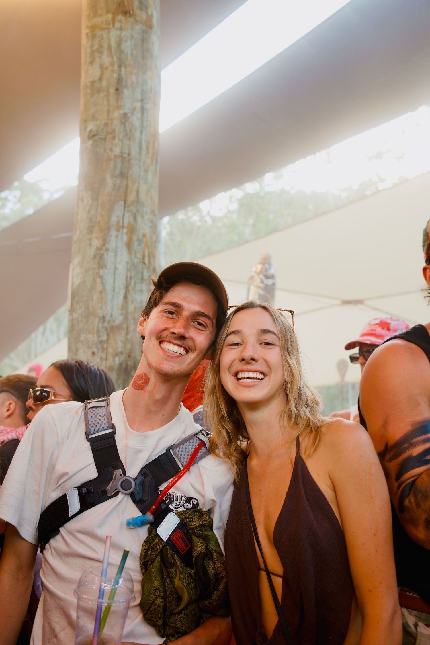 Smiling couple posing together at a sunny outdoor music festival under a shaded canopy.