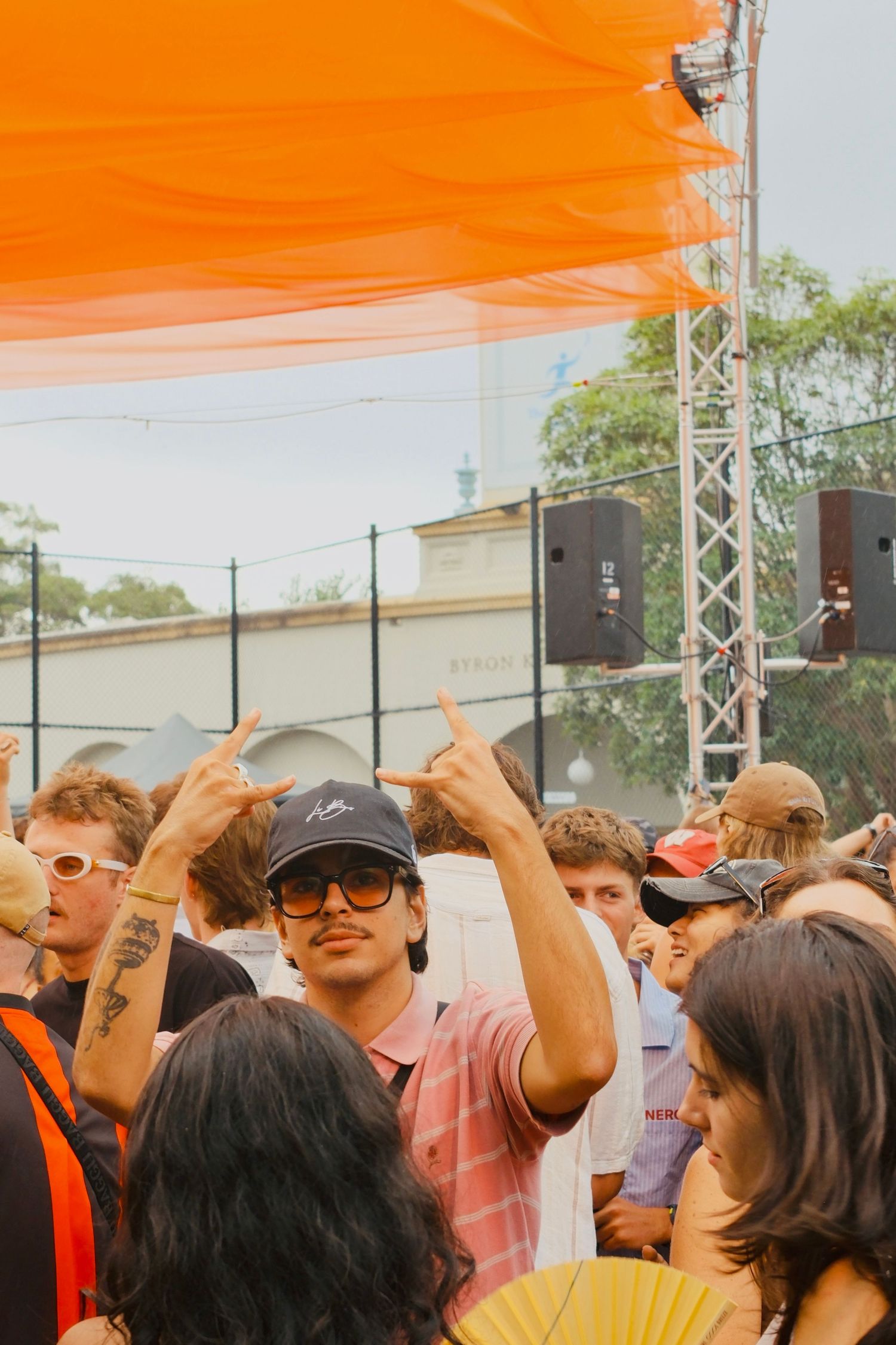 Young man in pink striped polo and black cap making rock on hand gesture in outdoor festival crowd.