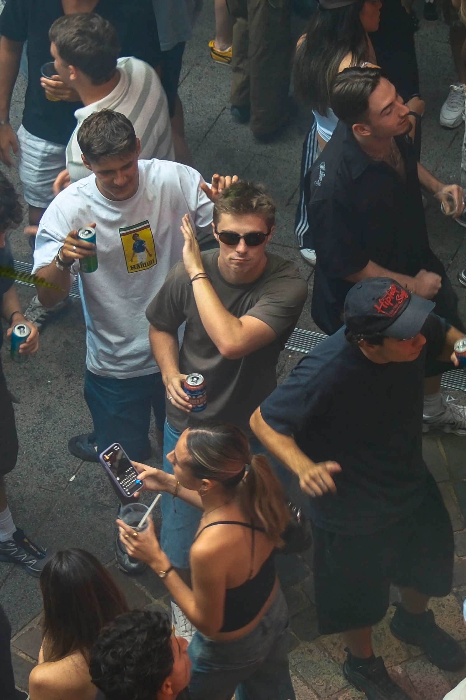 Crowd of young adults dancing and socializing outdoors at a street party, holding drinks and phones.