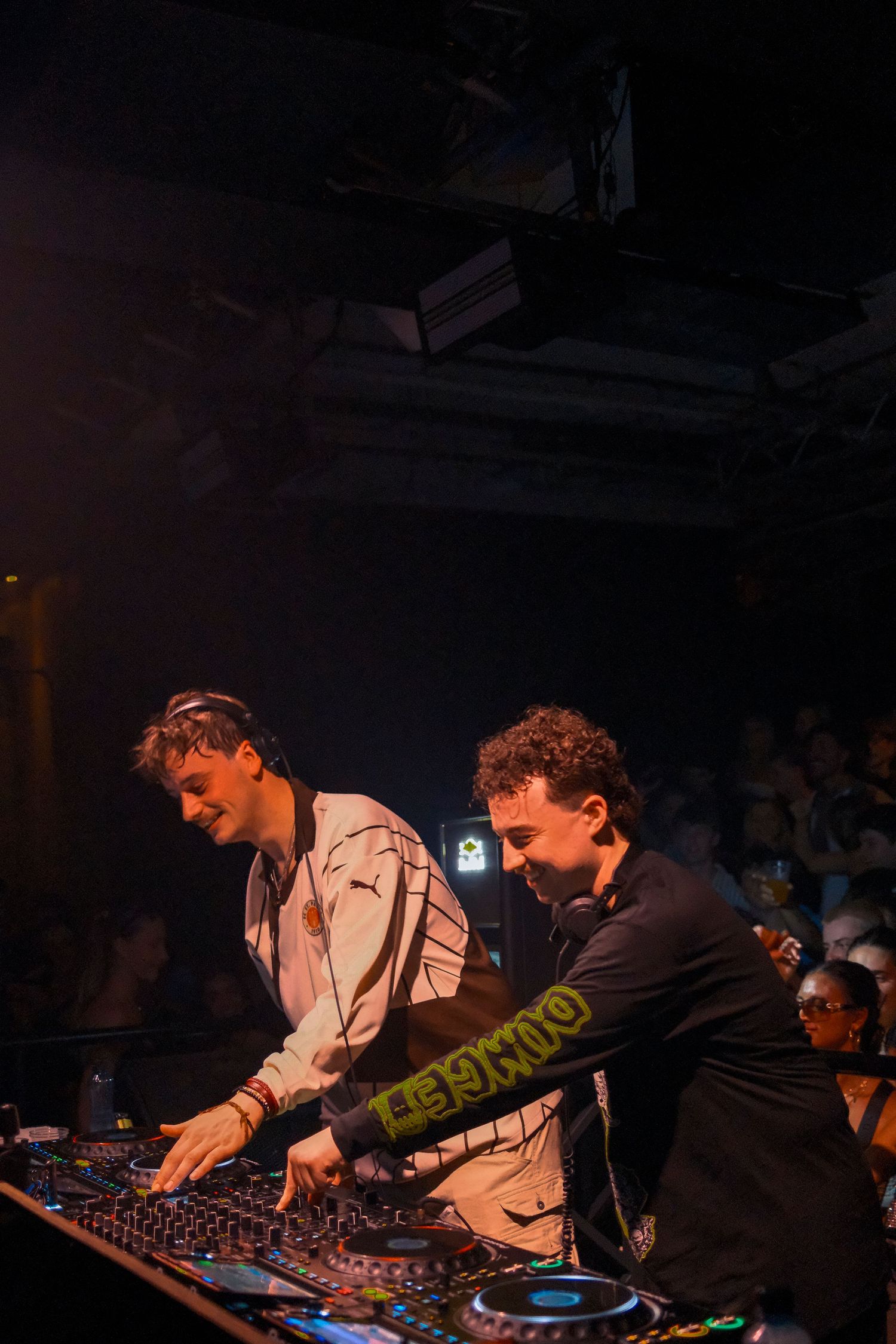 Two DJs performing at a nightclub, operating DJ equipment under dramatic stage lighting.