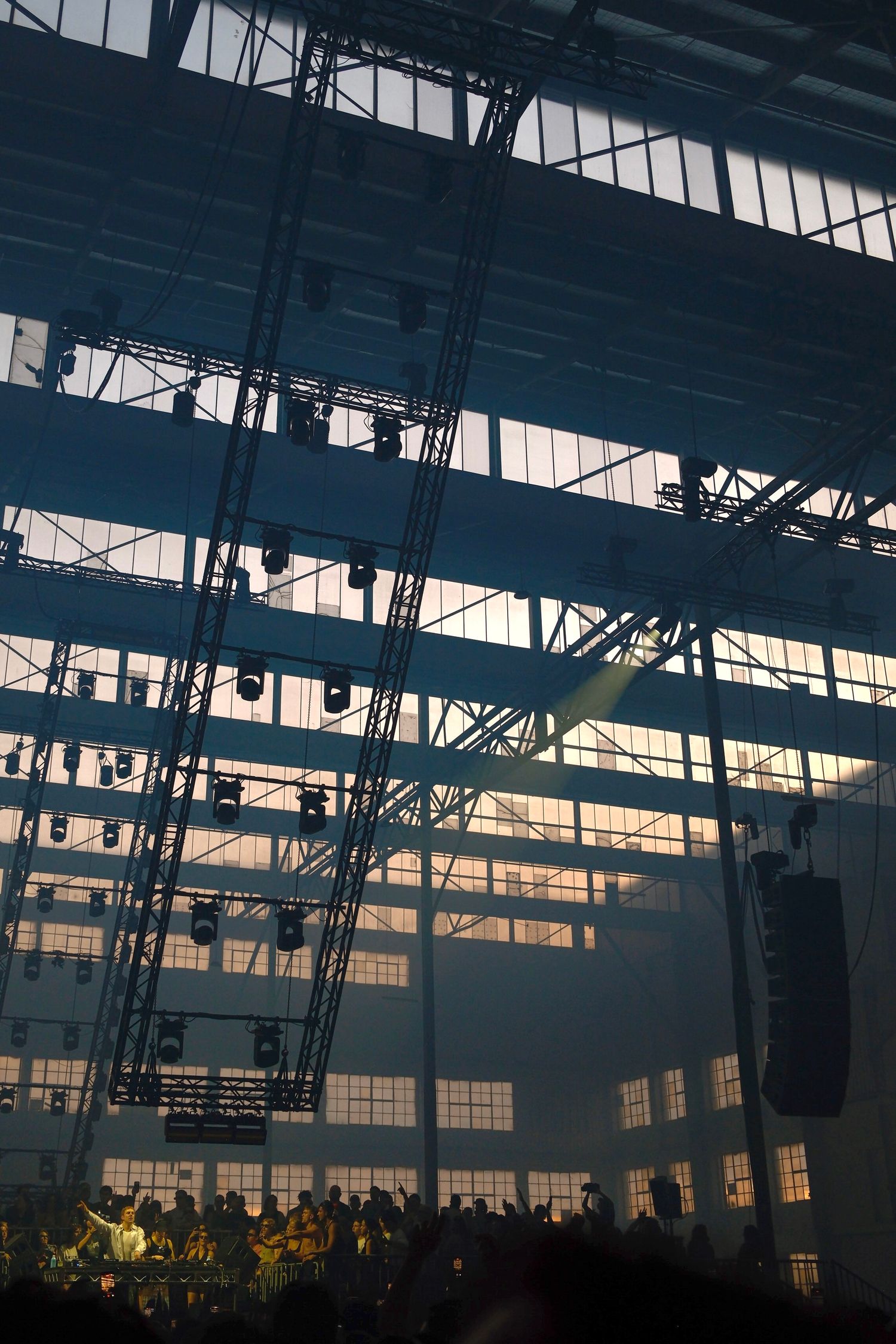 Large industrial warehouse concert venue with dramatic stage lighting and silhouetted crowd.