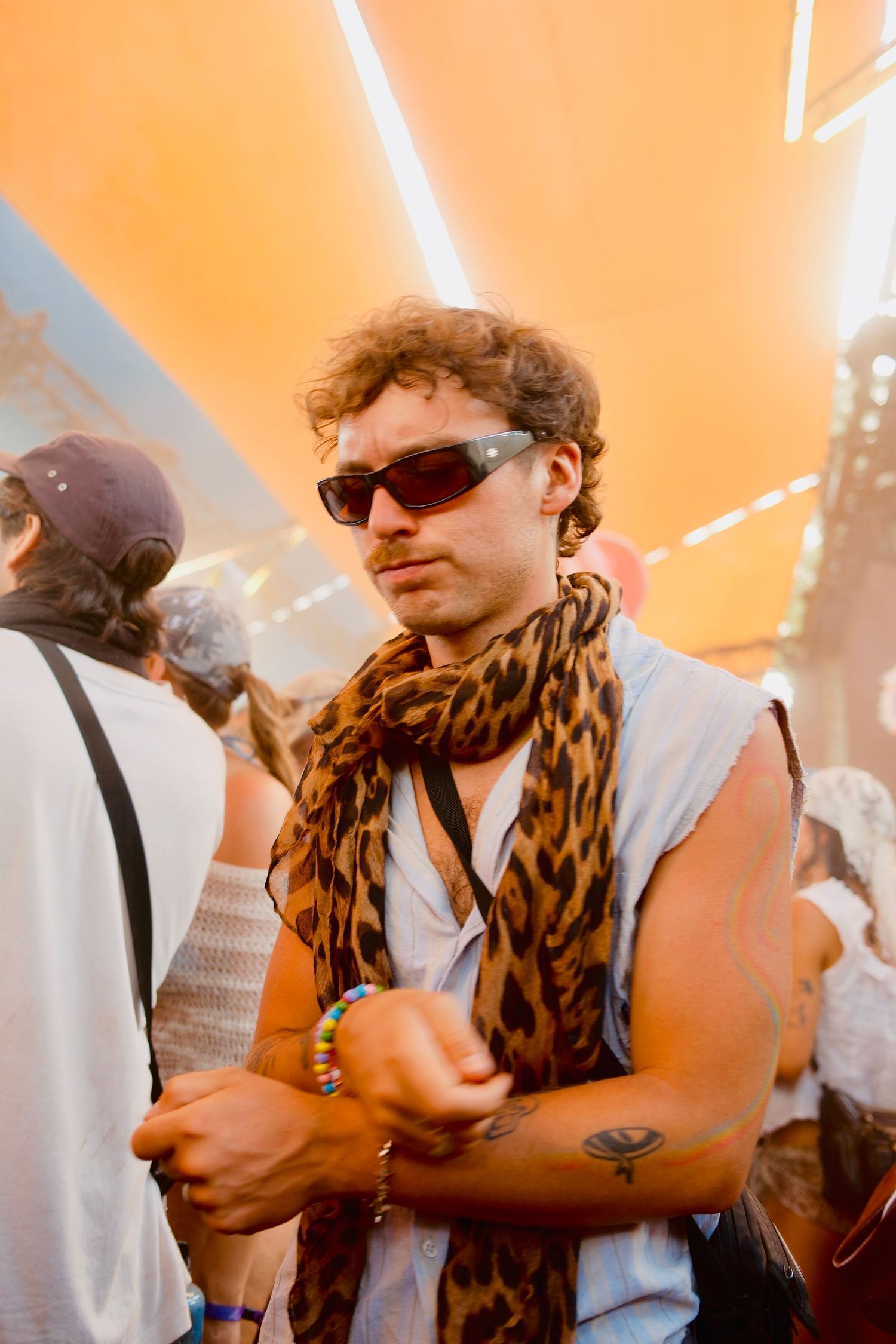Curly-haired man in sunglasses and leopard print scarf at a sunny outdoor festival.