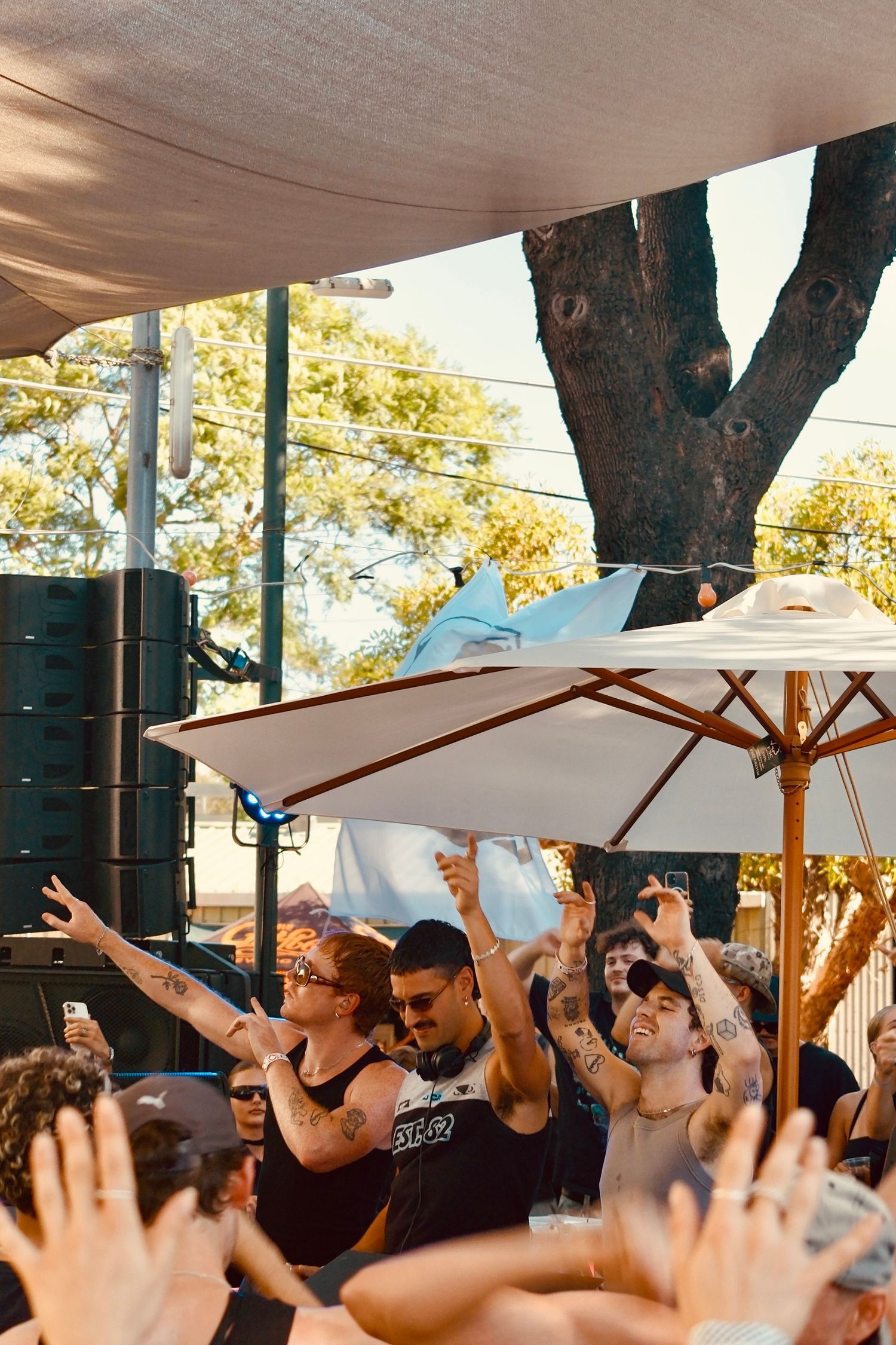 Energetic crowd with raised hands dancing at an outdoor music festival under a large patio umbrella.