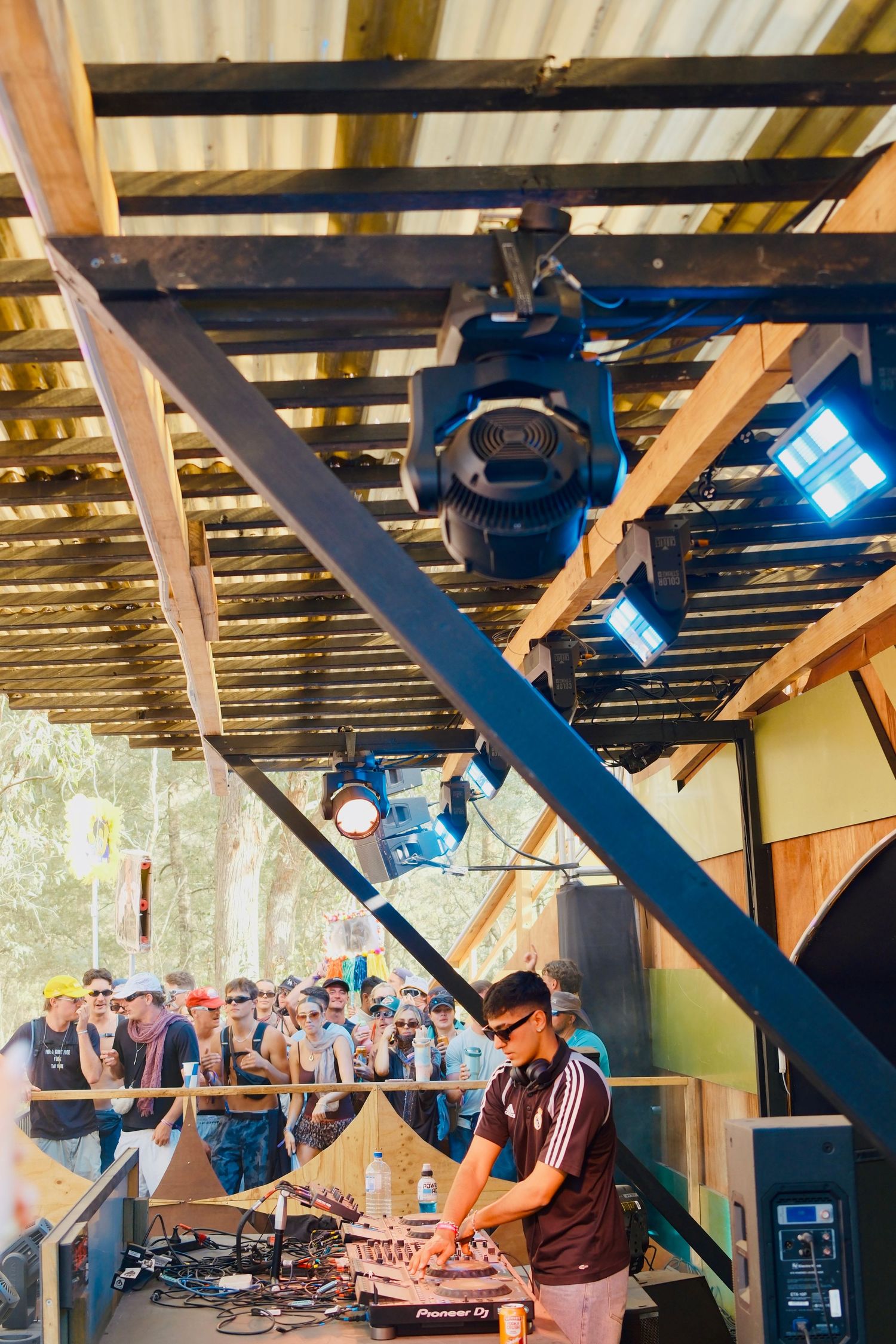 DJ performing on Pioneer decks under a rustic wooden stage roof with festival crowd watching outdoors.