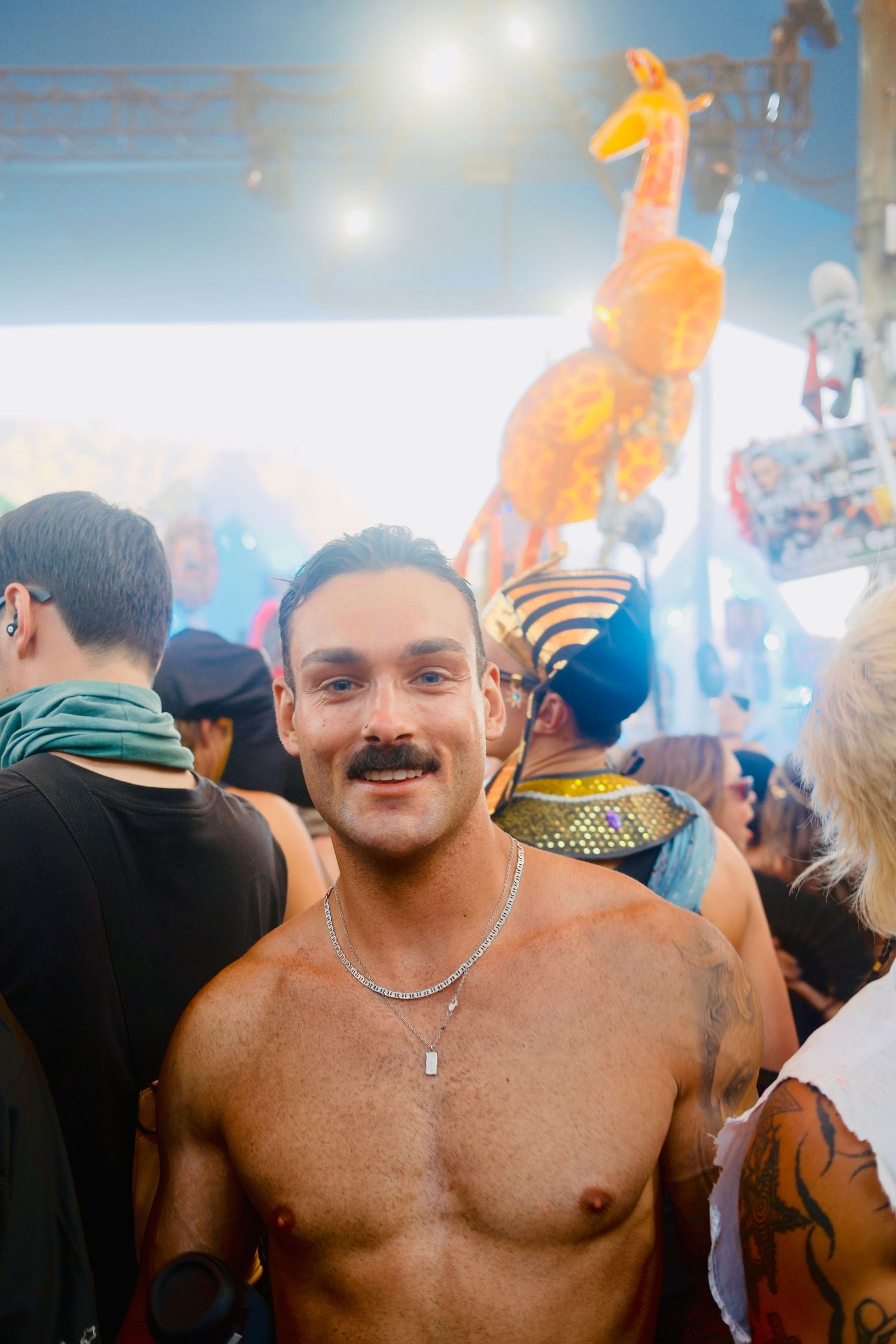 Shirtless man with mustache smiling at a lively outdoor music festival with giraffe decor.