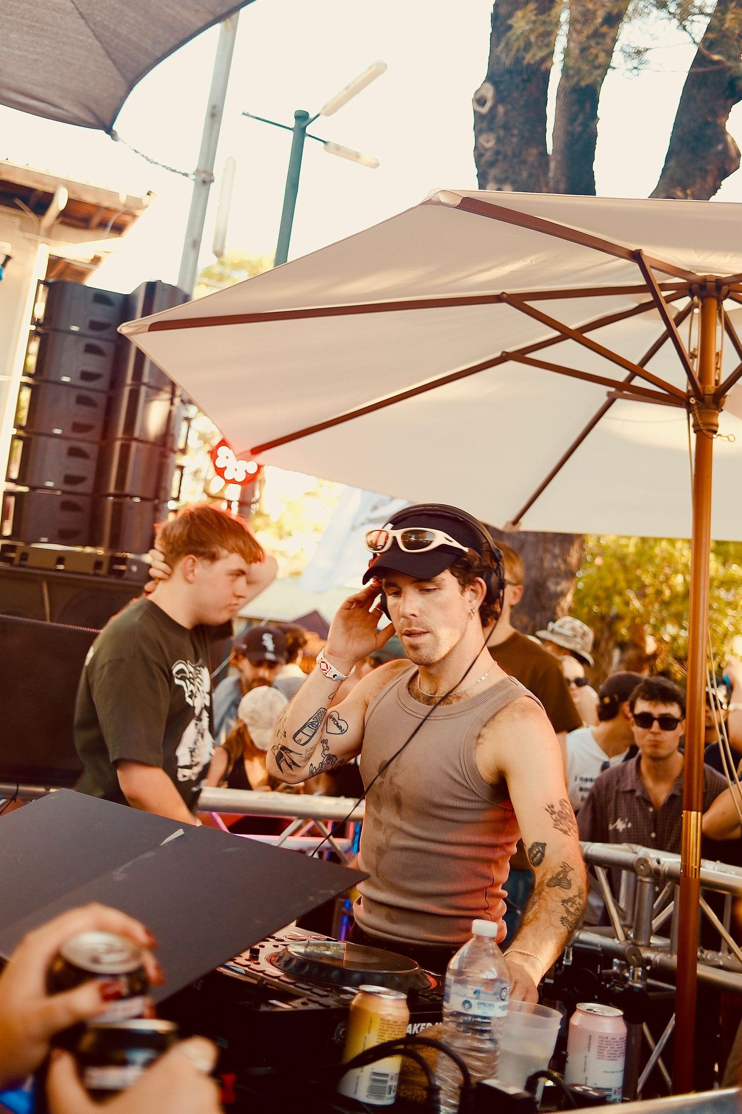 Female DJ with goggles and cap performs at a sunlit outdoor music festival stage.