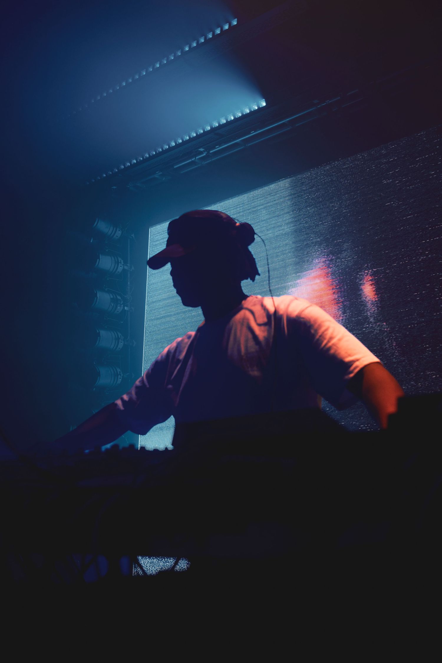 Silhouette of a DJ wearing a cap performing at a nightclub with blue and pink neon lighting.